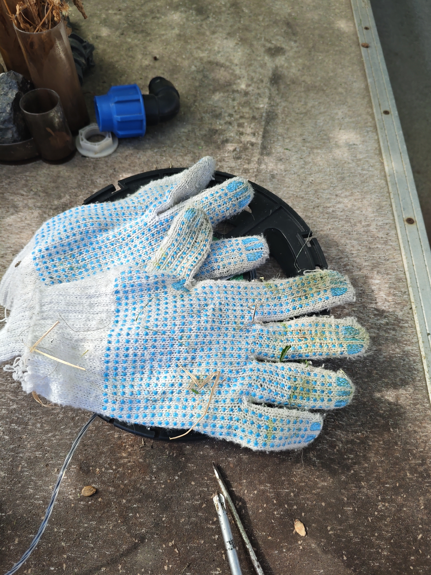 A pair of white and blue dotted work gloves lies on a concrete surface next to some plumbing fittings and tools. The gloves appear to be made of cotton with rubberized dots for grip. One fitting is blue and black, with a white threaded washer nearby. Two metal tools, possibly screwdrivers, are visible in the foreground. The concrete surface is mottled and appears to be outdoors, perhaps in a yard or workshop area. There are some dried plant materials scattered on the gloves and the ground. The lighting suggests it is daytime, with sunlight casting shadows. The scene is static and appears to be an outdoor storage or work area.