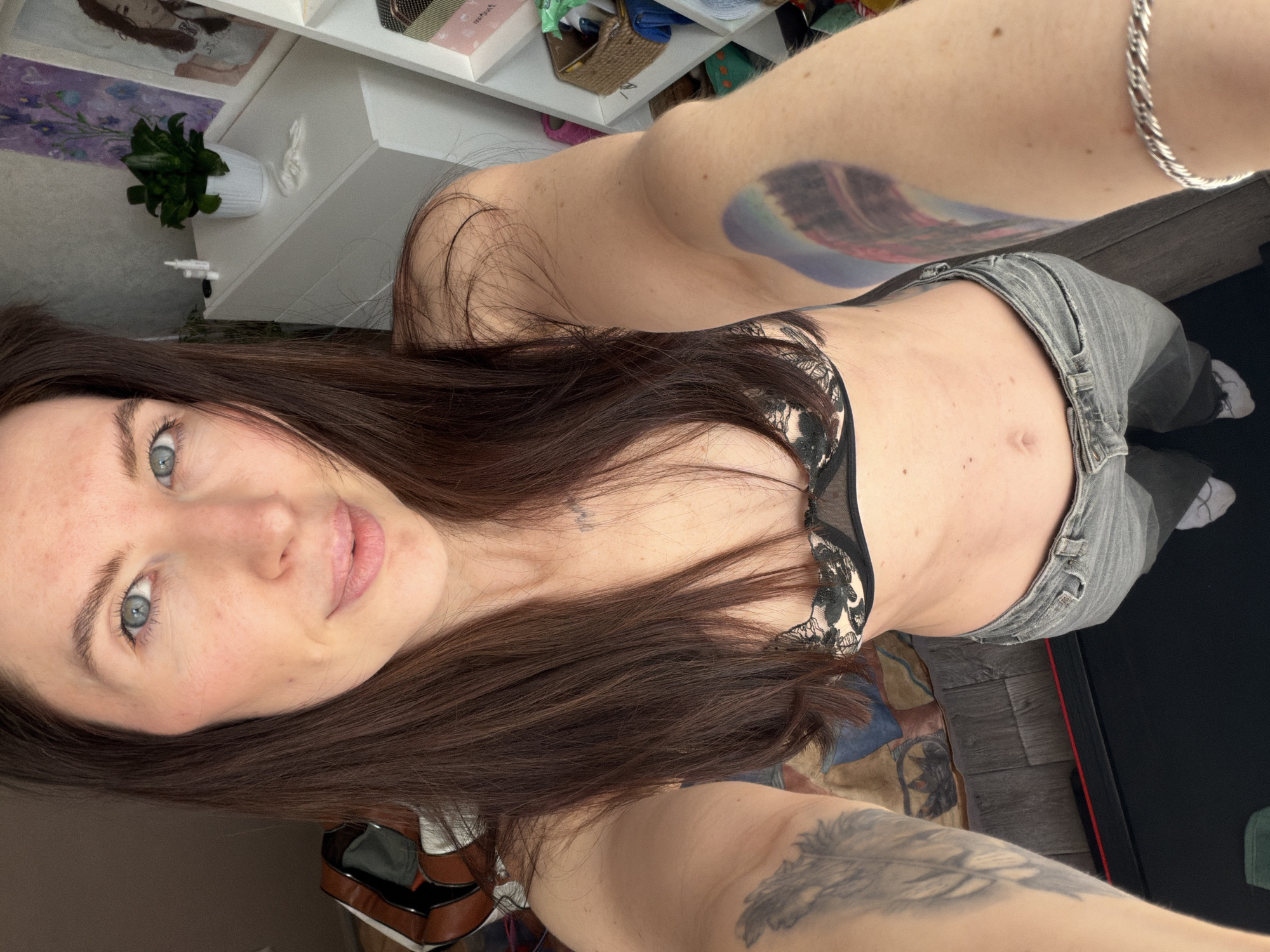 A woman is taking a selfie in what appears to be an indoor setting. She has long brown hair and blue eyes. She is wearing a black lace bra and gray jeans. There is a silver bracelet on her arm. She has tattoos on both arms. A white shelf with various items is visible in the background, along with a plant in a white pot. The location in the city of Perm, Russia, cannot be determined from the image.