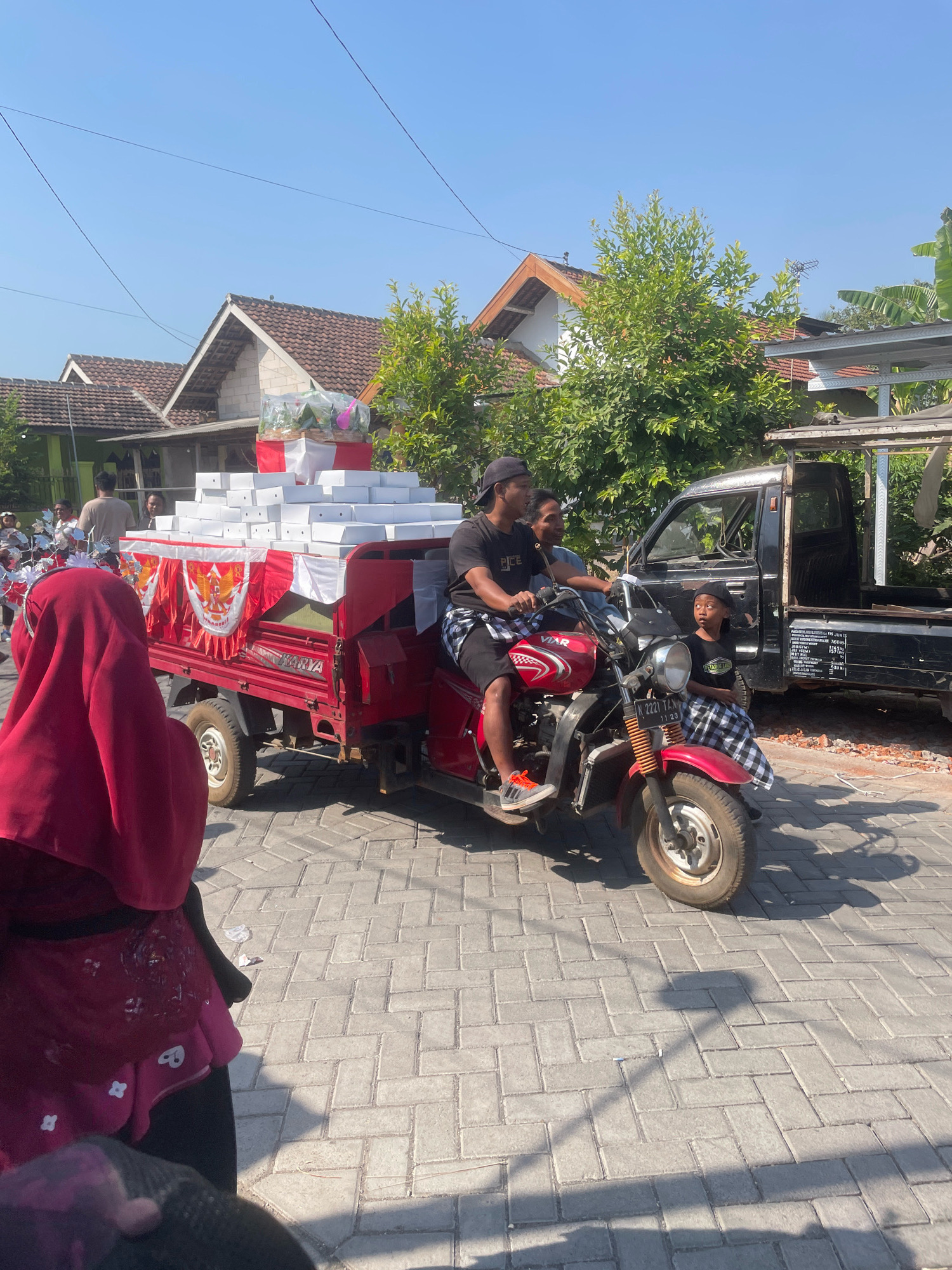 In the city of Kebon Waris, Indonesia, a red three-wheeled motor vehicle is loaded with numerous white boxes, potentially for a delivery or celebration. Two young men are seated on the vehicle, the driver wearing a black baseball cap and a black t-shirt with white lettering, and the passenger in a light blue shirt. A young child, also wearing a black baseball cap and a black and white checkered sarong, is seated on the front of the vehicle, looking to the left. The motor vehicle is adorned with red and white banners featuring an emblem. In the background, houses with tiled roofs and trees are visible under a clear blue sky. A person wearing a red headscarf and dark clothing is partially visible on the left side of the image, facing away from the camera. The vehicle is parked on a paved street.