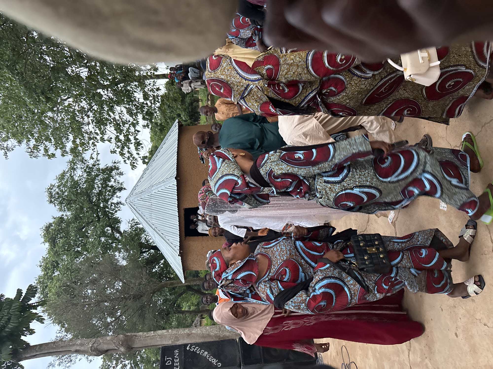 In a village setting outside of Jos, Nigeria, several individuals are gathered near a small, corrugated metal-roofed building. The ground appears to be dirt. On the left side of the image, a large black speaker is visible with "DJ ZEENR" and a phone number printed on it. A woman in a burgundy dress and headscarf stands to the right of the speaker, looking forward. In the center and right of the image, several women are dressed in colorful, patterned traditional clothing, with some wearing headscarves that match their outfits. One woman in the foreground is wearing a patterned dress and headscarf, holding a black handbag. Another woman next to her wears a similar outfit and holds a white handbag. A group of men are visible in the background, some standing near the building and others sitting. Trees and foliage are present in the background, and the sky is overcast. The overall atmosphere suggests a community gathering or celebration.