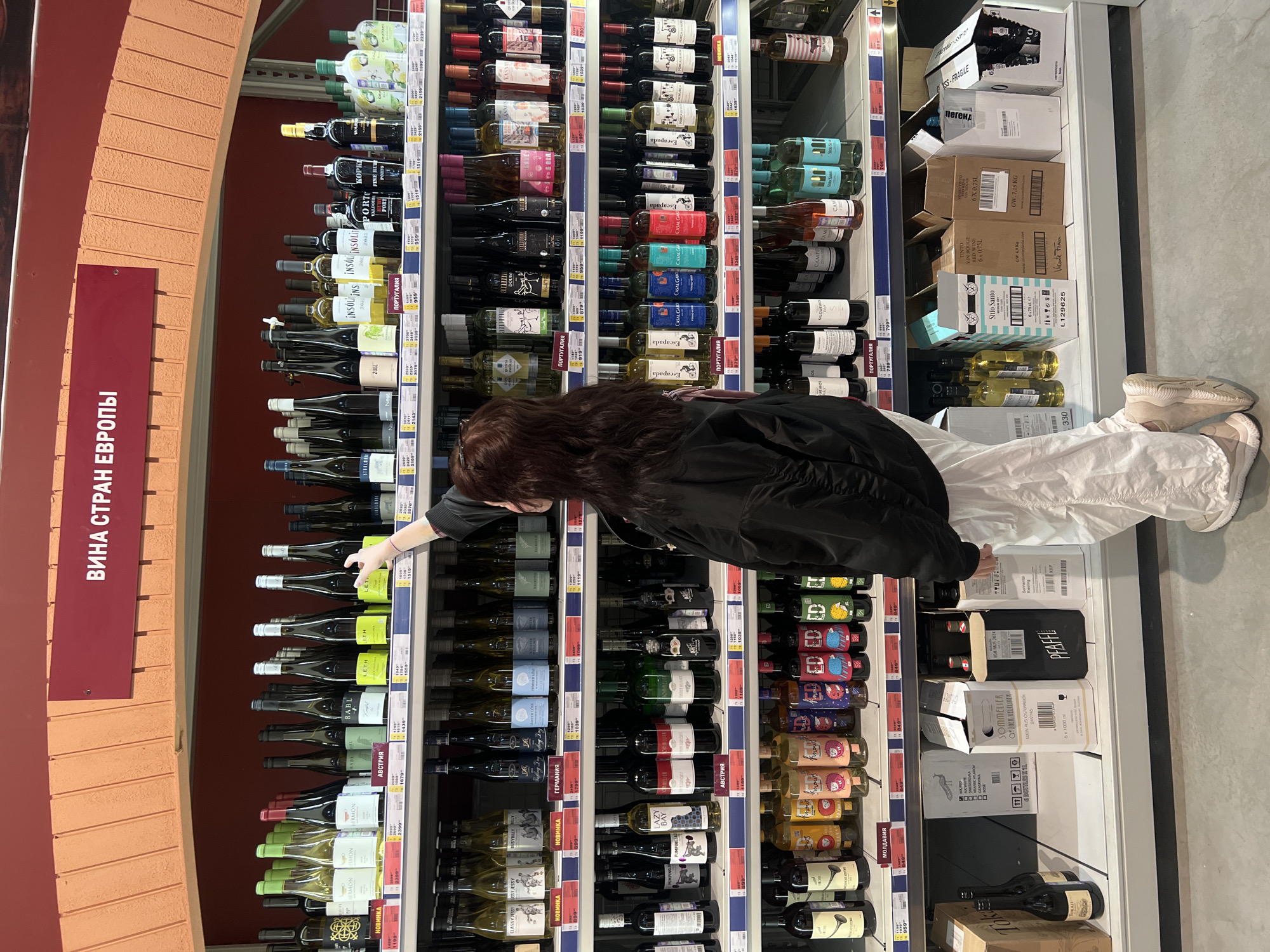 A woman with long, dark hair and wearing white pants and a black jacket is standing in front of a shelf stocked with wine bottles in a store in Izhevsk, Russia. She is reaching towards a bottle of white wine on the shelf. The shelves are labeled with the names of the countries from which the wines originate, including Portugal, Germany, and Austria. A sign on the left side of the frame reads "ВИНА СТРАН ЕВРОПЫ" which translates to "Wines of European Countries." The store is well-lit and the floor appears to be a light gray color. Additional wine and boxes of wine are visible on the shelf to the right.