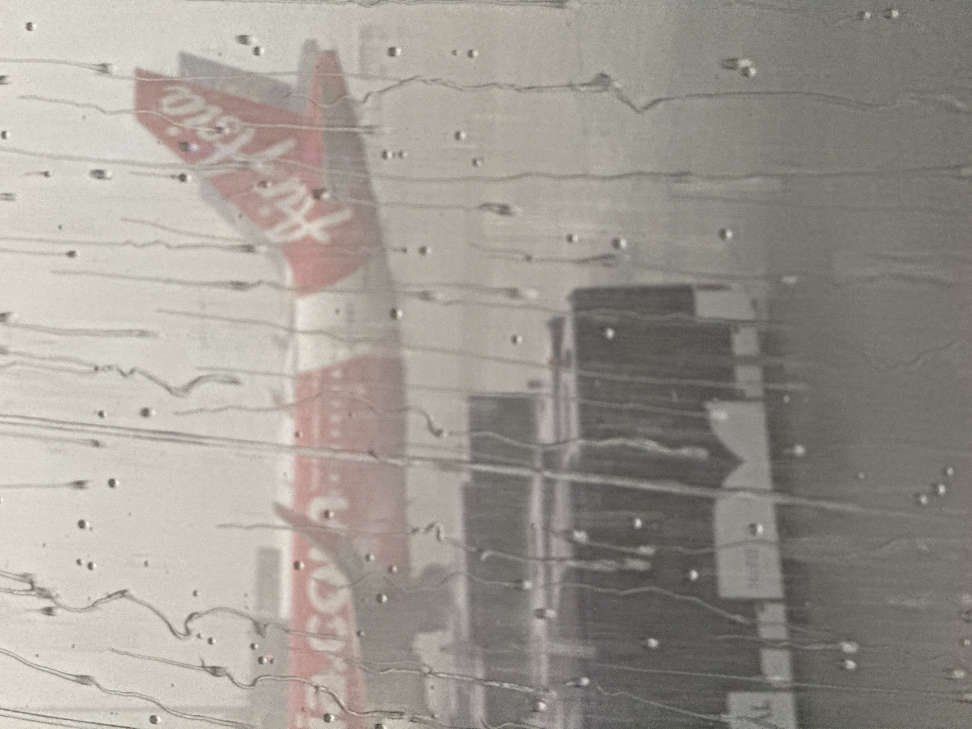 An AirAsia airplane is visible through a window covered in water streaks and droplets. The airplane is predominantly red with white lettering that reads "Air Asia" on its tail. In the foreground, partially obscured by the water, is a section of a bus or service vehicle, which appears to be dark purple with white markings. The scene suggests an airport environment, possibly during rainy weather, given the water on the window. The overall image is blurry and desaturated due to the rain and the viewing angle. The location is described as Nong Prue Subdistrict, Thailand.