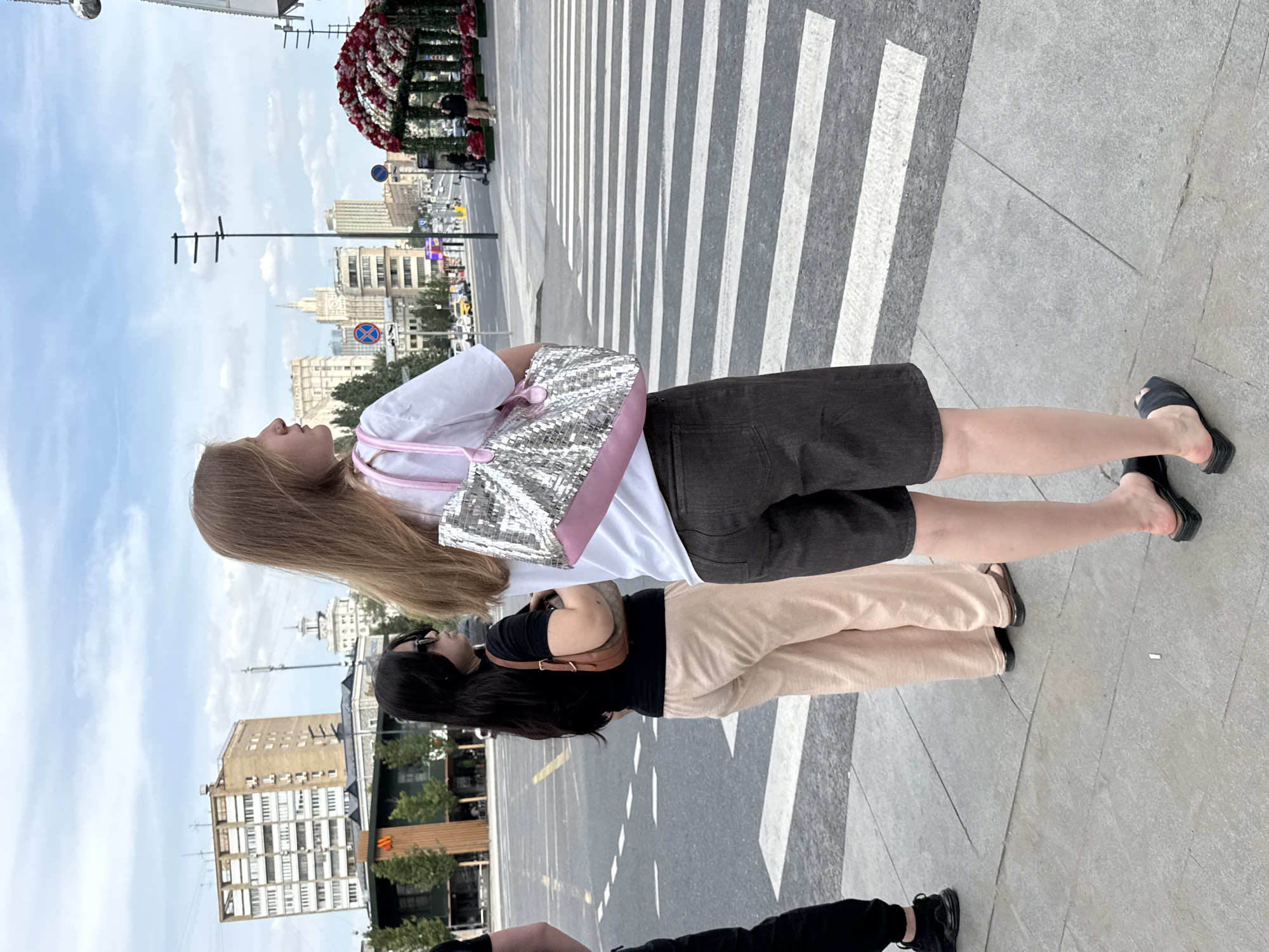 The image shows three individuals on a city sidewalk next to a crosswalk. The person in the foreground, a young woman with long blonde hair, is facing away from the camera, wearing a white t-shirt, dark shorts, and black slides. She is carrying a silver sequined handbag with pink trim. To her left, another woman with long dark hair and sunglasses is also facing away from the camera. She is wearing a black t-shirt and light-colored pants, carrying a brown shoulder bag. A third person, only partially visible on the far left, is wearing black pants and black sneakers. In the background, there is a city street with traffic, buildings, streetlights, and a decorative floral archway. The overall environment appears to be an urban setting in Moscow, Russia, likely a pedestrian area with a significant crosswalk. The sky is overcast with clouds.