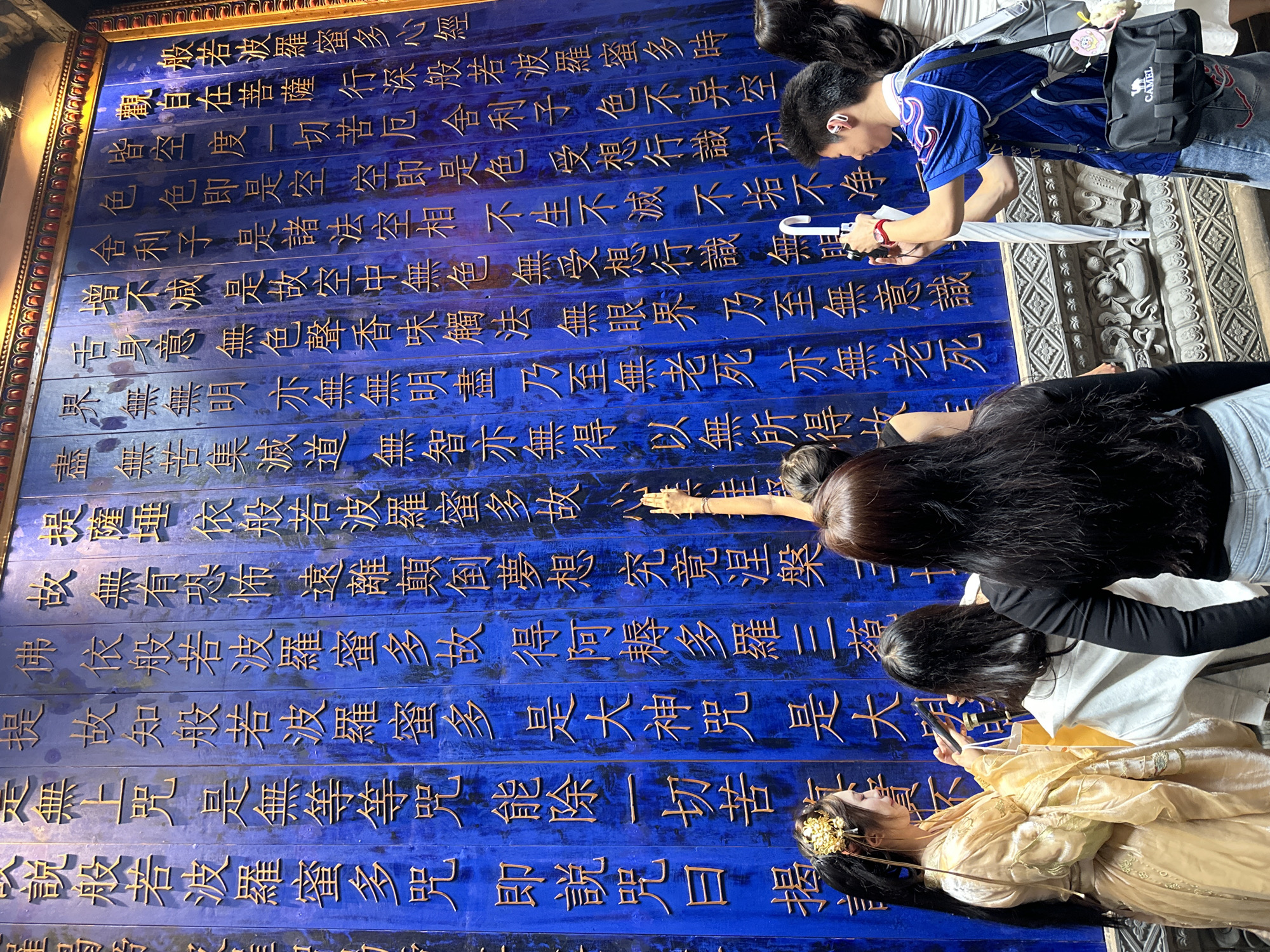 The image shows a group of people interacting with a large wooden wall engraved with golden Chinese characters on a blue background. One person is taking a photo of the wall with a smartphone, while another person wearing a blue shirt and carrying a bag is holding an umbrella nearby. A woman in traditional Chinese clothing is posing for a photo. The wooden wall seems to be a display or exhibit of sorts, possibly found in Yuzhong District, China, featuring a classic Chinese Buddhist text, the Heart Sutra, written in traditional characters. The overall setting appears to be a tourist spot or a cultural heritage site, where visitors are engaging with and capturing the experience.