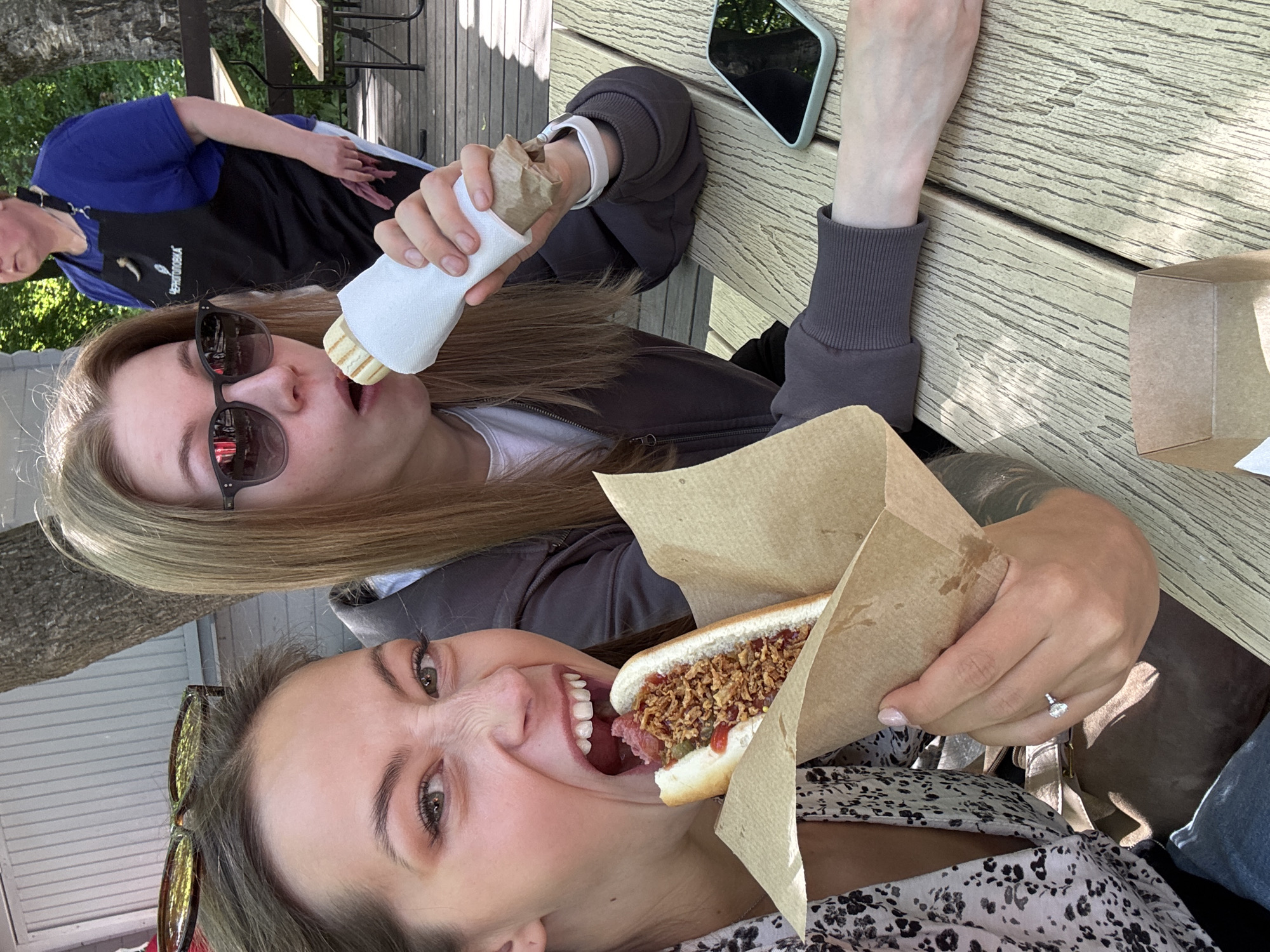 Two young women are captured in a bright, sunny outdoor setting, enjoying hot dogs at a wooden picnic table. In the foreground, one woman with long brown hair, wearing a patterned top and sunglasses pushed up on her head, takes a large, happy bite of her hot dog, which is topped with what appears to be crispy fried onions and red sauce. A simple ring is visible on her left hand.
Behind her, another woman with blonde hair, wearing sunglasses and a dark grey hoodie, holds a wrapped hot dog in one hand and a white, conical condiment dispenser in the other, aiming it towards her hot dog. Her expression is partially obscured by her large sunglasses, but she appears relaxed.
In the background, a person in a blue shirt and a black apron, possibly a food vendor, is visible, partially obscured. Text on the apron is mirrored, appearing as "dooF teertS", likely indicating "Street Food". A light-colored smartphone is placed on the wooden table beside the women. The overall scene suggests a casual and enjoyable outdoor meal, characteristic of a street food experience or an informal eatery in Saint Petersburg, Russia during the daytime.