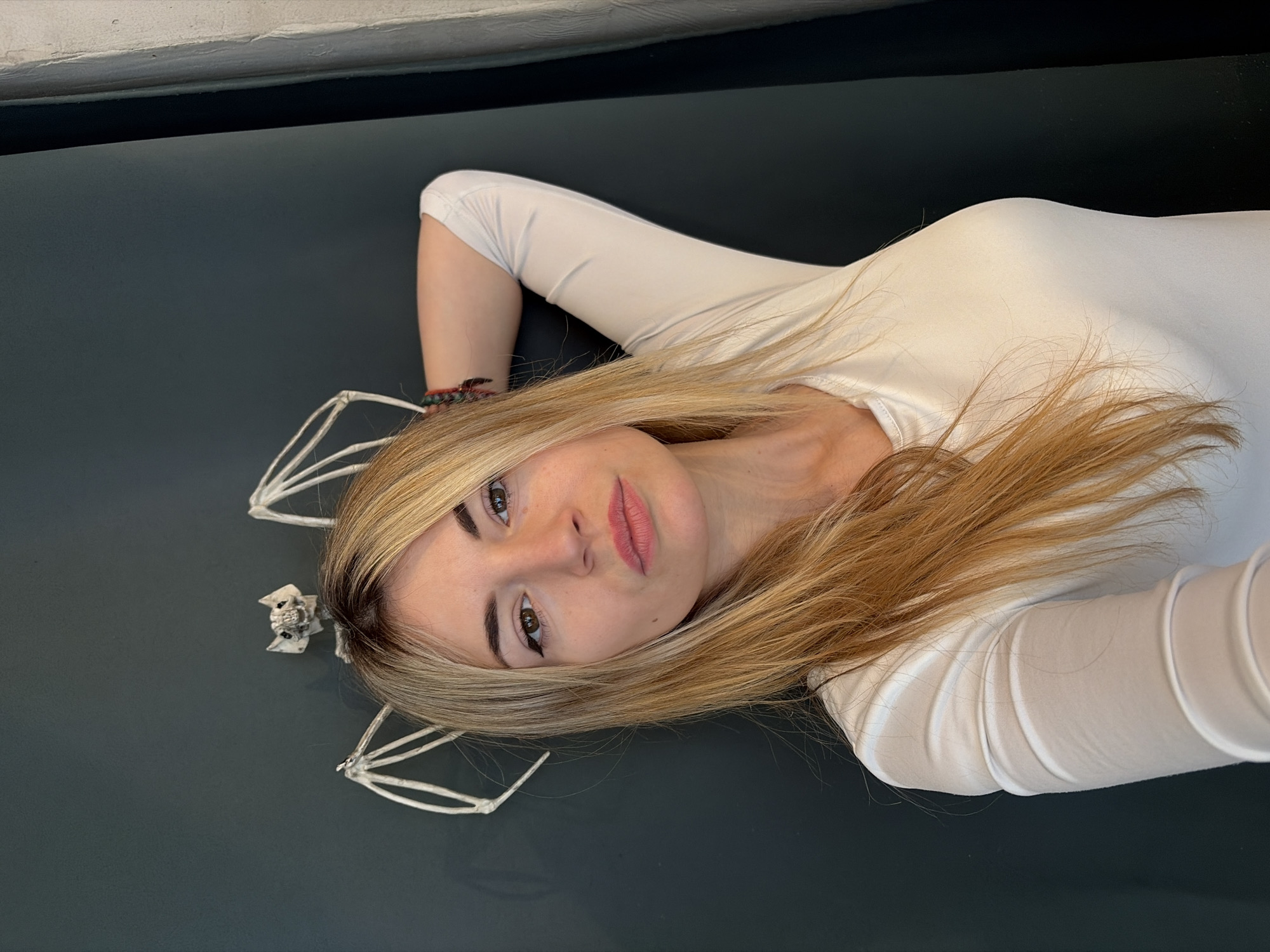 A young woman lies on her back on a dark surface, wearing a white long-sleeved shirt. Her blonde hair is spread around her head. To her left, closer to the top of the image, there are skeletal decorations. She appears to be looking directly at the camera. The setting is a dark surface with a black and white border. Without other details to reference, I cannot verify the location in the city of Budva, Montenegro.