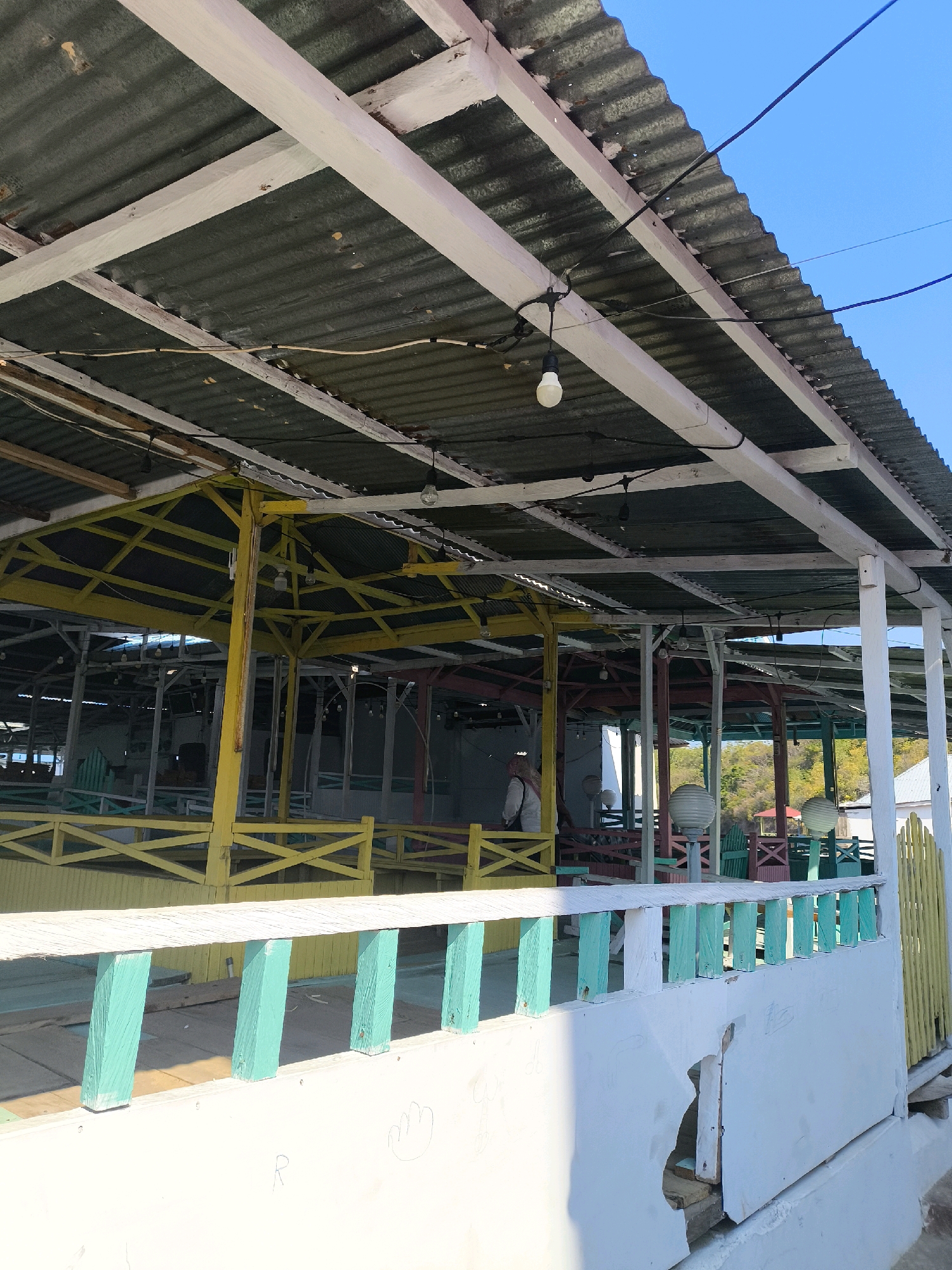 The image shows an outdoor seating area, likely a cafe or restaurant, with a corrugated metal roof supported by a wooden frame. The structure is painted in various bright colors, including yellow, green, and white, with some areas showing signs of wear and tear. A person wearing a white jacket and dark pants is partially visible in the background. The area appears to be situated near a body of water, possibly the sea or a lake, given the presence of a clear blue sky and distant trees on a hill. This scene is likely located in the city of Tilamuta, Indonesia, known for its coastal scenery and vibrant local establishments.