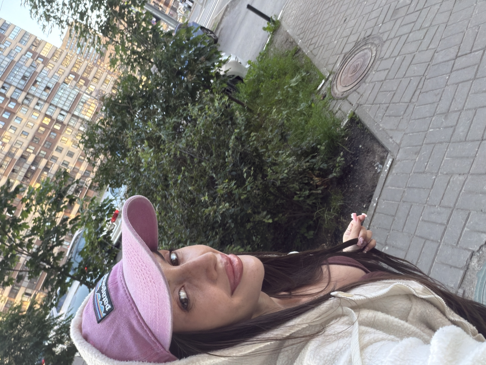 A young woman is taking a selfie outdoors in a city environment. She is wearing a pink baseball cap with a Patagonia logo and a cream-colored hooded jacket. She has long, dark hair and is smiling, with her face turned towards the camera. Behind her, there are green trees and bushes, a multi-story building with many windows, and a paved sidewalk with a manhole cover. The scene suggests a residential area, possibly in Saint Petersburg, Russia, given the architectural style of the building. The lighting indicates it might be late afternoon or early evening.