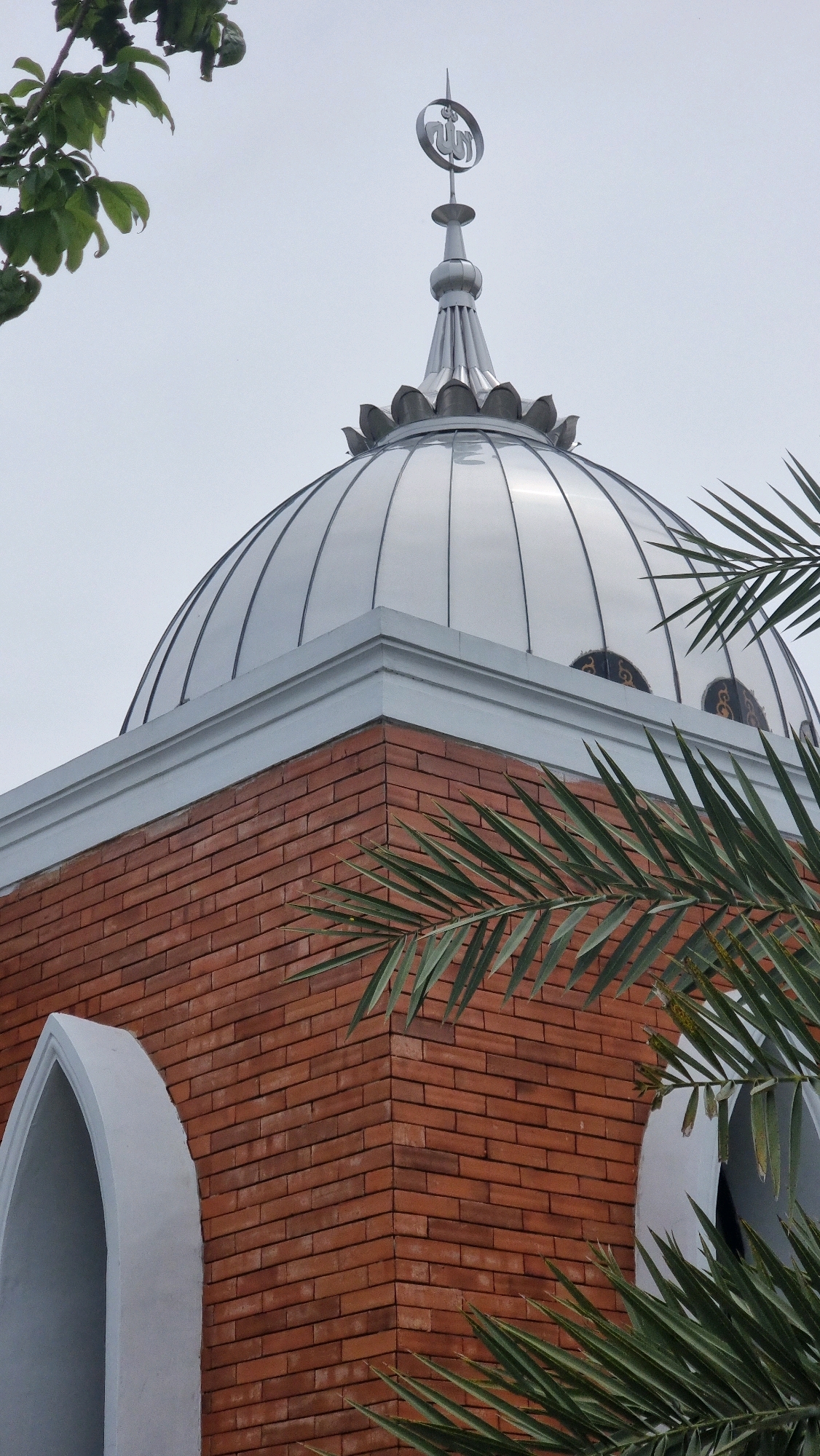 A medium shot captures a mosque dome and its apex, possibly in Lhoksukon, Indonesia, under an overcast sky. The dome is a polished silver, reflecting the light with smooth, vertical segments. It is topped by a spire adorned with a crescent moon and Arabic calligraphy, likely the word "Allah." The dome sits atop a brick structure, a corner of which is prominently featured. The bricks are reddish-brown and laid in a standard running bond pattern. A white, decorative band runs along the top of the brickwork, supporting the dome. On the left side of the image, a pointed archway, painted white, is visible. Palm fronds from a nearby tree partially obscure the view of the dome and the brick structure, with some leaves in the foreground and others in the background, suggesting it is an outdoor scene.