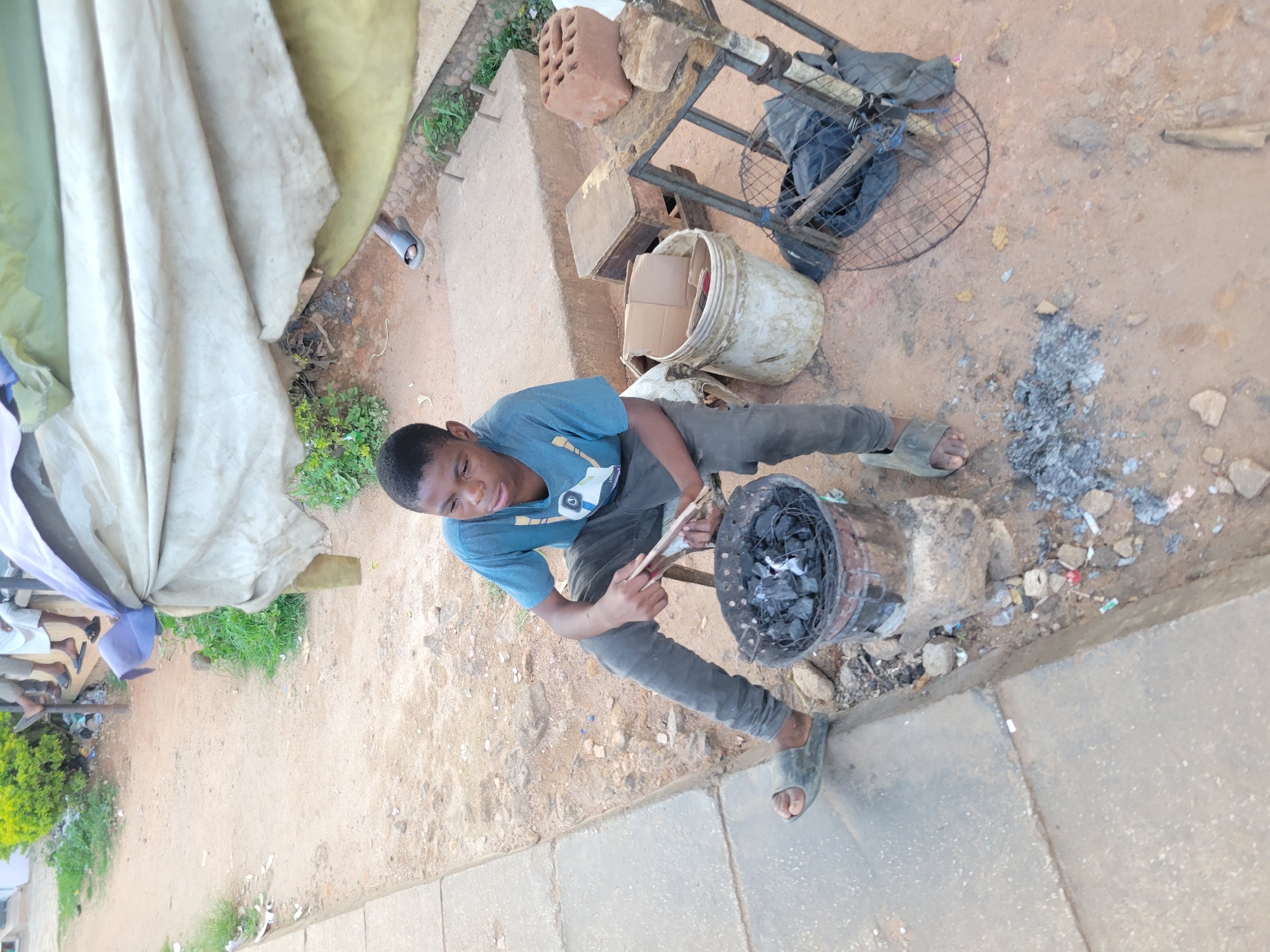 The image depicts a young boy in Jos, Nigeria, tending to a charcoal brazier. He is wearing a blue t-shirt and dark pants, with grey sandals on his feet. The boy is actively engaged in stoking the coals with a stick, suggesting he is preparing to cook or is in the process of cooking.
The setting appears to be an outdoor, informal marketplace or roadside area. Various objects are scattered around, including bricks, a metal frame, a plastic bucket with cardboard inside, and a discarded grill. The ground is dirt and gravel, with some scattered debris and patches of greenery. A large, light-colored tarp or tent is visible on the left side of the frame, possibly providing shade. The presence of ash and discarded items hints at a working or frequently used area.
The lighting suggests it is daytime, though the weather is not explicitly discernible. The boy's facial expression is focused on his task, showing concentration. The overall scene conveys a sense of everyday life and labor in a busy, informal urban environment. There is no visible text in the image.