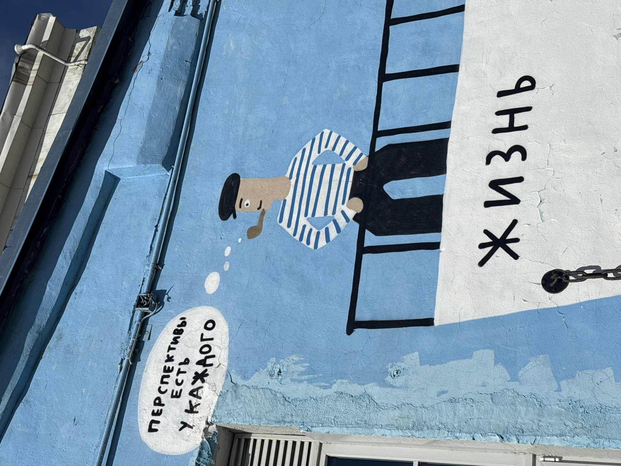 A mural in Saint Petersburg, Russia, depicts a stylized sailor in a striped shirt and beret, smoking a pipe while climbing a ladder towards the Russian word "Жизнь" (Life). A thought bubble near the sailor contains the phrase "Перспективы есть у каждого" (Everyone has prospects). The mural is painted on a light blue exterior wall with a window and various architectural details. The building appears aged, with cracked and peeling paint.