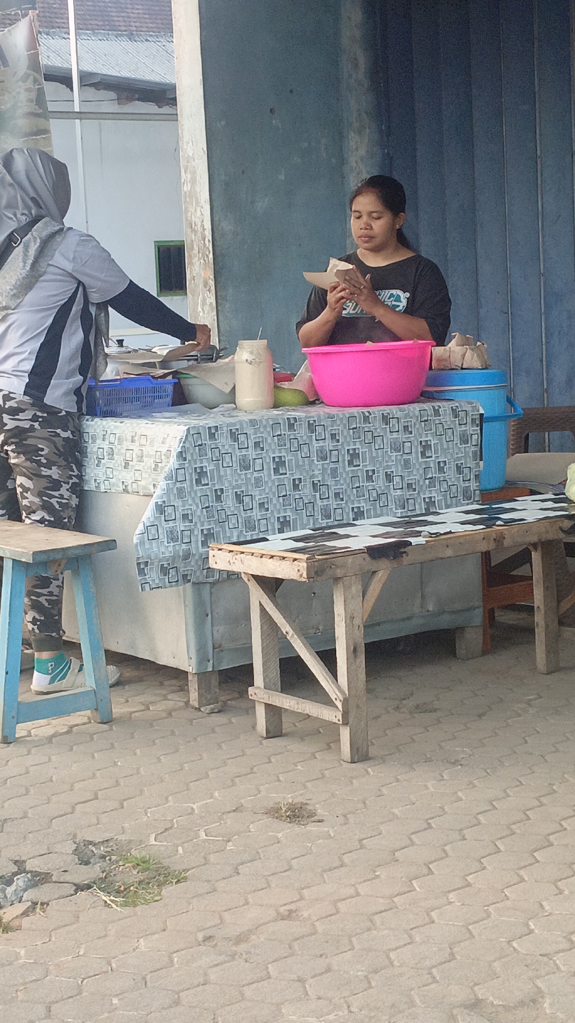 A woman stands behind a food stall in Srono, Indonesia, holding a piece of paper in her hands. She is wearing a black t-shirt. To her left, another person in a gray hijab and a black and white striped shirt is leaning over the stall, appearing to handle something on the surface. The stall is covered with a patterned tablecloth and displays a pink bowl, a glass jar, and a blue basket. A blue cooler sits to the right of the stall. In front of the stall, a wooden bench with a black and white checkered surface is visible. The ground is paved with hexagonal gray bricks. The background shows a building with a corrugated metal roof and a blue slatted wall.