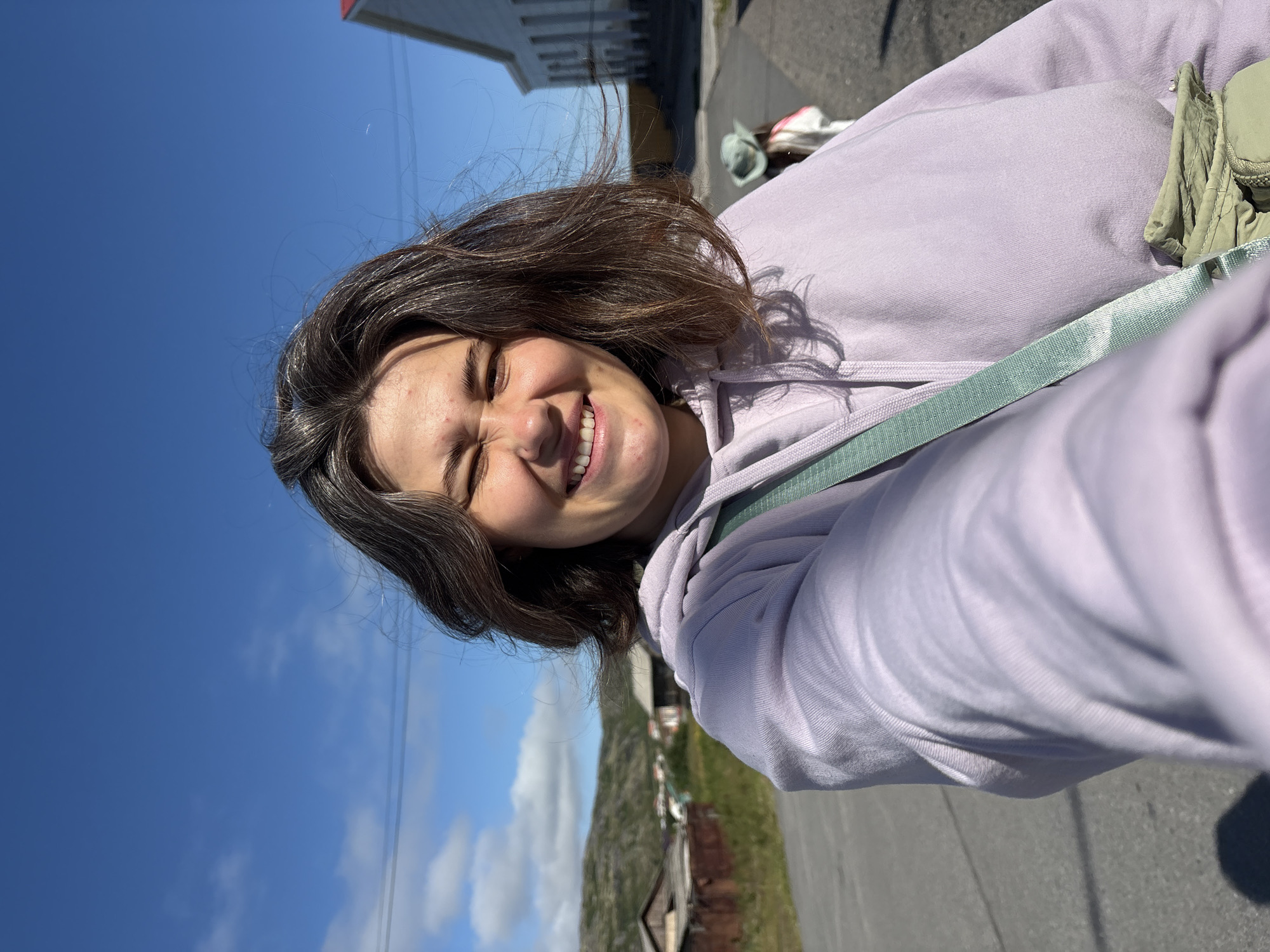 A young woman with shoulder-length brown hair is taking a selfie outdoors on a sunny day. She is wearing a light purple hoodie and a light green crossbody bag. Her eyes are squinted, and she has a wide smile. In the background, there is a paved road, some rustic wooden buildings, a green hillside, and a blue sky with scattered clouds. To the right, a person wearing a light-colored jacket and a hat is walking away from the camera. The architecture and the general landscape suggest a setting in a northern climate, consistent with Teriberka, Russia.