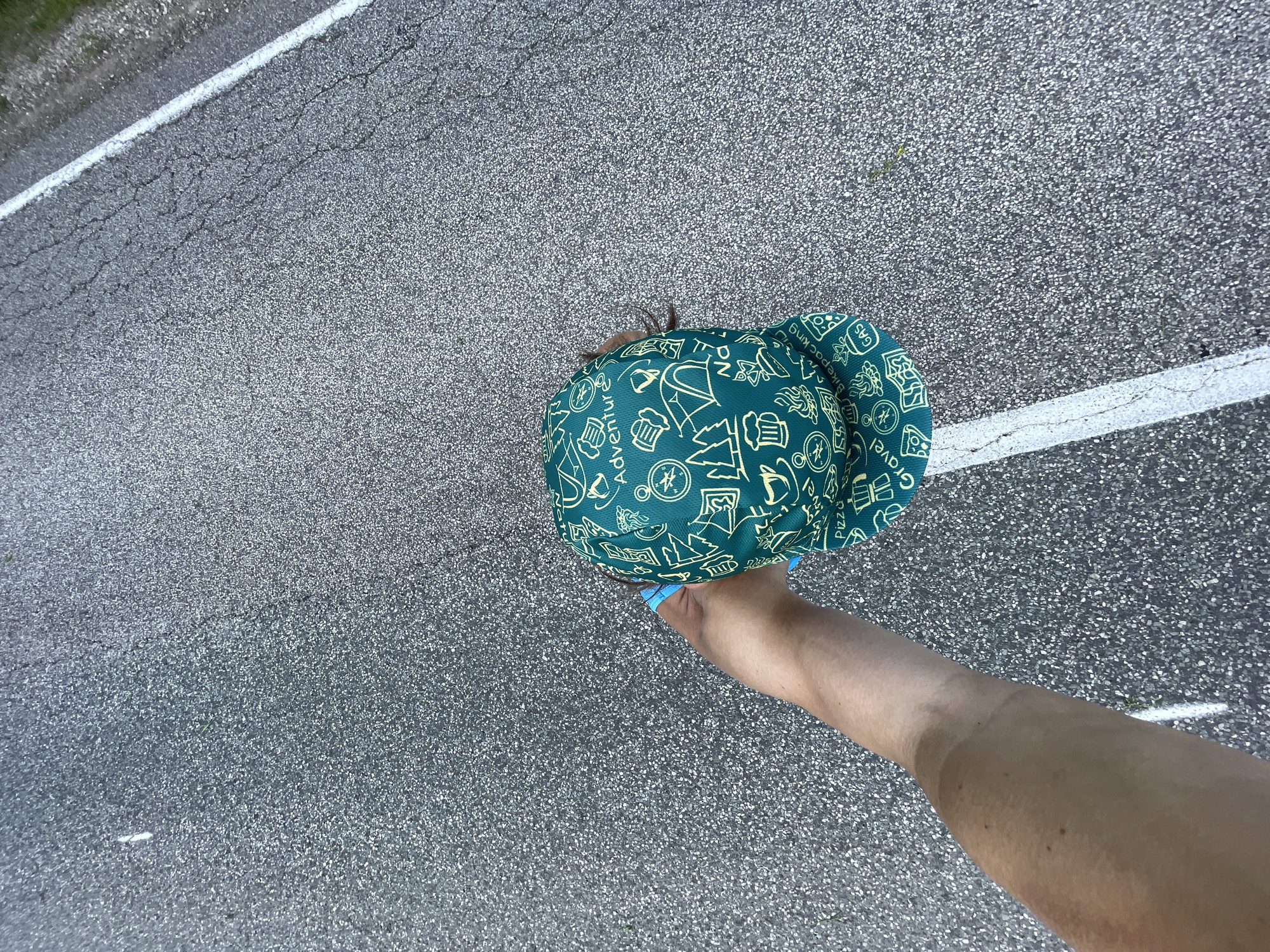 An overhead, close-up shot shows a person's arm and hand holding a green baseball cap. The cap has a pattern of yellow icons, including tents, trees, and compasses, and the word "Adventure." The background is asphalt with a white line marking a road. The scene is outdoors, and it appears to be daytime.