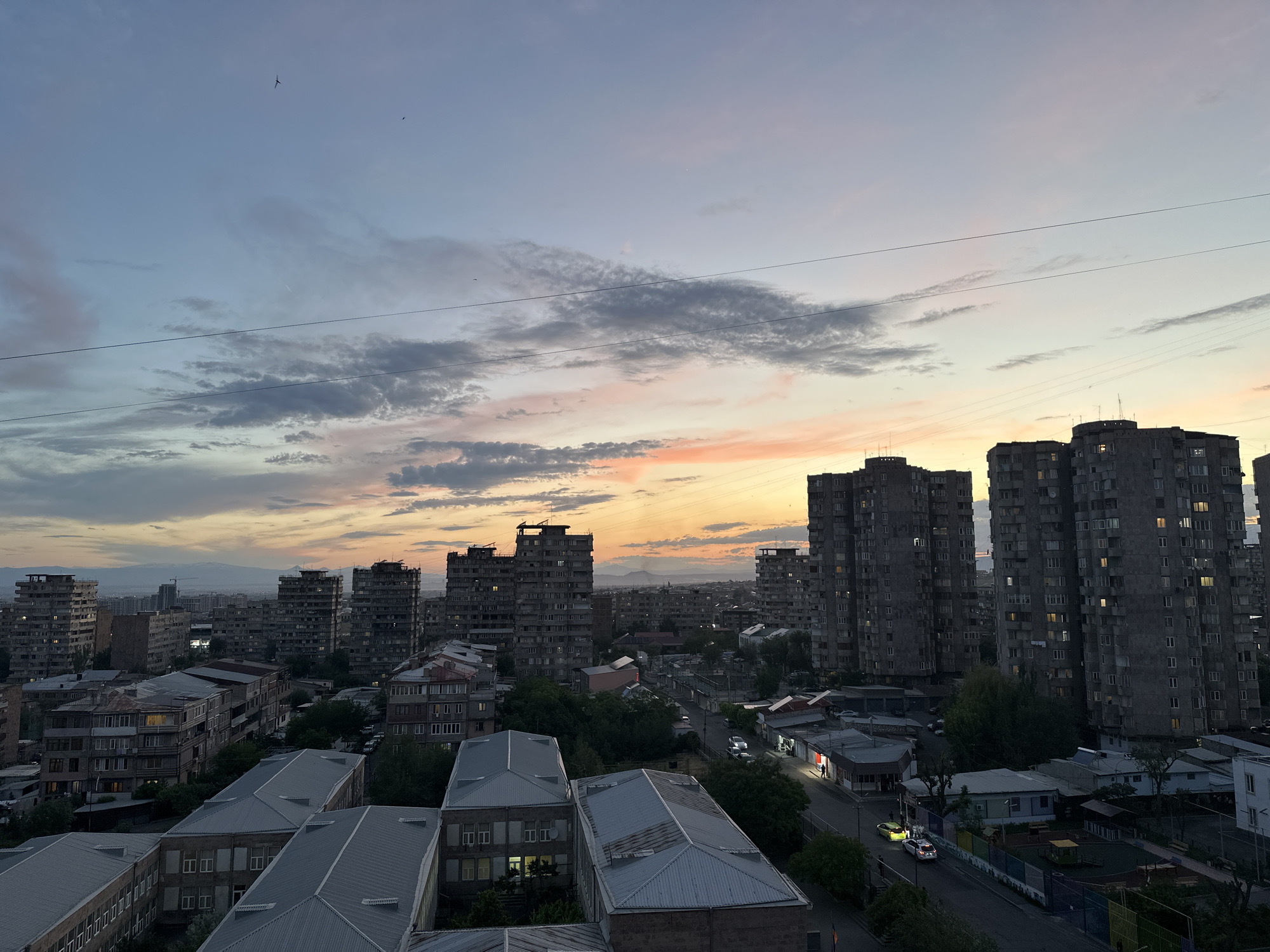 This elevated panoramic image captures a dense urban residential district in Yerevan, Armenia, during twilight. The sky transitions from a pale blue at the zenith through layers of light grey and pink clouds, culminating in a bright orange and yellow glow at the western horizon, indicative of a sunset. Multiple thin black power lines stretch horizontally across the upper and middle portions of the sky. Several small, dark bird silhouettes are visible against the upper sky.
The cityscape is dominated by numerous multi-story, grey, brutalist-style concrete apartment buildings, characteristic of Soviet-era architecture, distributed across the mid-ground and background. Many windows in these large blocks are illuminated, indicating evening activity. Below these towers, a network of smaller residential and commercial structures with varying roof types, including several prominent grey corrugated metal pitched roofs, fills the foreground and mid-ground.
A winding street, illuminated by streetlights and the headlights of moving vehicles, cuts through the lower mid-ground. At least two light-colored cars are visible on the street. Several storefronts along the street are brightly lit, with one featuring a distinct red glow. Two individuals can be observed near a brightly lit commercial establishment on the right side of the street. Adjacent to the street, a children's playground with colorful equipment is partially visible. In the distant background, mountains are silhouetted against the lighter sky, with some peaks displaying lighter patches, possibly snow or bare rock.