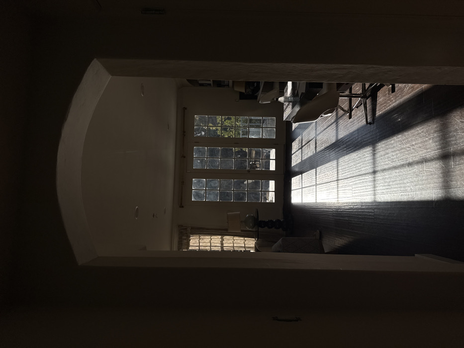 The image depicts the interior of a residential space in Los Angeles, California, United States. The vantage point is from a dimly lit hallway, looking through an arched doorway into a brightly sunlit room.
The room features dark, wide-plank hardwood flooring, across which strong, rectangular shafts of natural light from windows or glass doors are cast, creating distinct patterns of light and shadow. At the far end of the room, multiple glass-paned French doors or large windows, divided into numerous square sections, overlook an outdoor area with visible green foliage.
To the left, a window with light-colored curtains is present. Below it, a small, dark-colored round table with an ornate base holds a table lamp with a light-colored shade. A portion of a patterned armchair or sofa is partially visible to the immediate left of the table.
To the right, a dark desk or table is situated, accompanied by two white, modern-style swivel chairs with chrome bases. A clear water bottle or container is visible on the desk surface. Behind the desk, a built-in bookshelf filled with books is discernible. The walls of the room are light-colored, and the ceiling features recessed lighting fixtures. No individuals are present, and no actions or interactions are depicted.