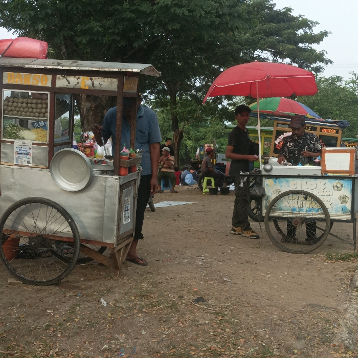 This image depicts an informal outdoor food vending area located within the Special Capital Region of Jakarta, Indonesia. The ground is unpaved dirt with scattered foliage and minor debris, extending into a background dominated by mature trees with dense green canopies.
In the left foreground, a stainless steel "Bakso" food cart with a wooden base is visible. Its front display section contains spherical food items (bakso balls), noodles, and other ingredients, with a price sign indicating "Rp 15.000". A vendor, clad in a light blue short-sleeved shirt, is partially visible behind the counter, appearing to prepare food; his head is out of frame. A large, circular metal plate rests on the counter, alongside a holder containing bottles of red and green condiments. The cart is mounted on two large bicycle wheels.
To the right, a second food cart, primarily white with blue accents, is positioned under a large red umbrella. A vendor, wearing a patterned short-sleeved shirt and a brown cap, stands behind this cart, engaged with a white rectangular container on the counter, likely holding ingredients for cold drinks or desserts. A young male in a dark t-shirt and camouflage pants stands between the two carts, facing the right-side vendor. This cart also features bicycle wheels and has a white, blank sign board affixed to its front right. A smaller, multi-colored umbrella is partially visible behind this cart.
In the background, beneath the trees, several individuals are present. Some are seated on the ground or on low stools, suggesting customers or people resting. A green plastic stool and a white sheet or tarp are also visible in this area. The scene is illuminated by diffused daylight.
