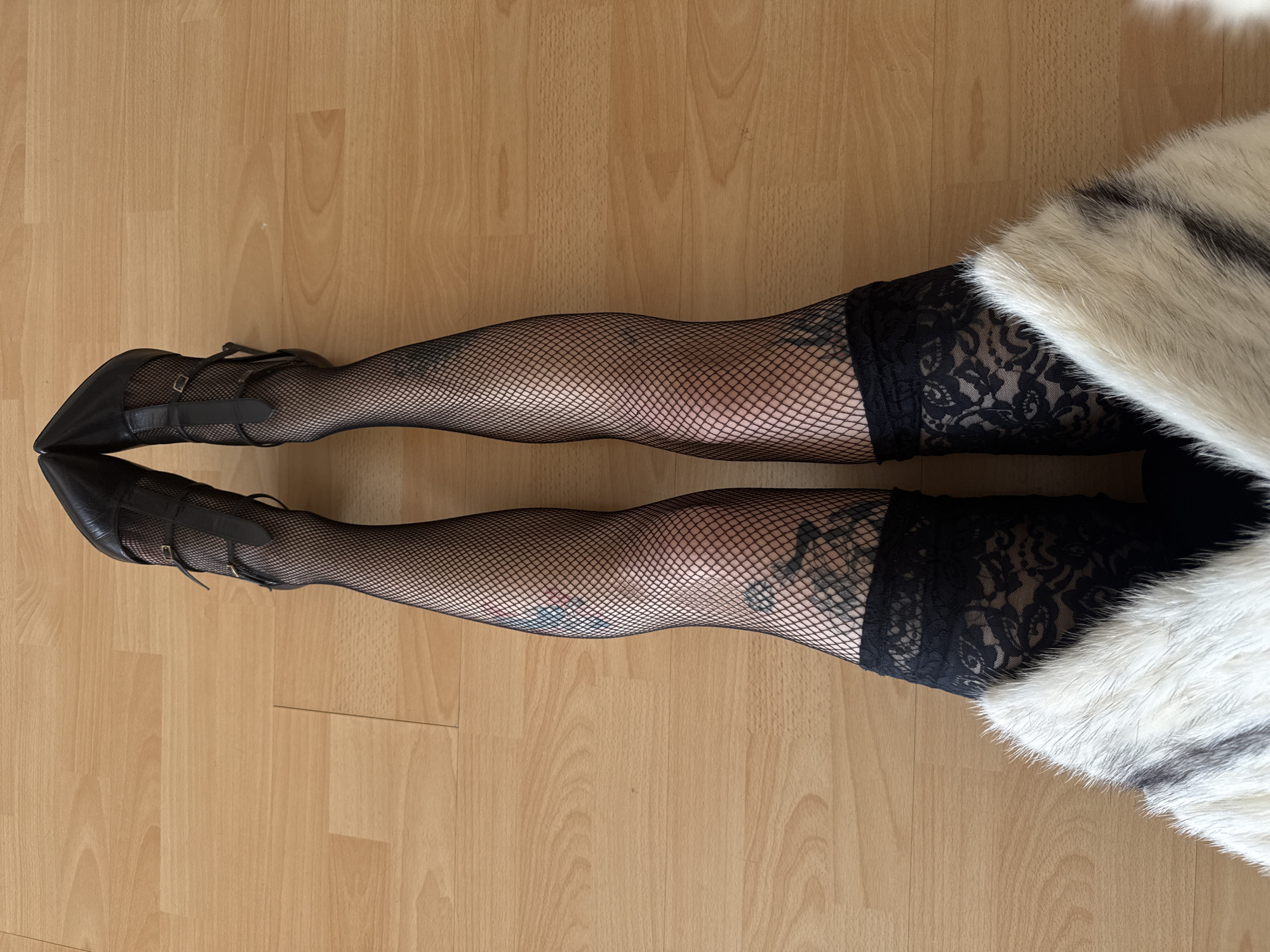 A person's legs, clad in black fishnet stockings with black lace thigh-high tops and black pointed-toe Mary Jane high heels, are captured resting on a light brown wooden floor. The legs are extended directly away from the viewer. A white and black fur or faux fur throw partially covers the upper thighs and hips on the right side of the frame. Visible tattoos are present on both legs underneath the fishnet material; one prominent dark tattoo is seen on the left thigh/knee area, and another dark, possibly abstract tattoo is on the right thigh/knee area. A smaller, possibly colored tattoo is also visible on the lower right leg, near the ankle. The setting appears to be indoors with bright, even lighting. There is no discernible activity or event taking place other than the pose itself. No text is visible within the image.