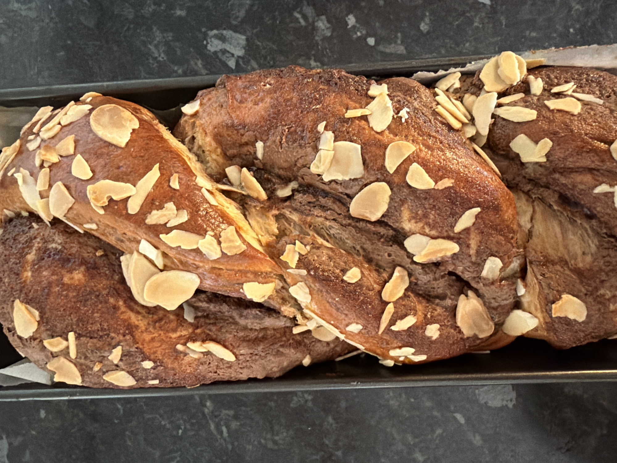 A freshly baked, almond-topped braided bread loaf sits in a dark metal baking pan. The loaf has a dark brown, slightly mottled crust and is generously sprinkled with almond slices. The pan rests on a dark, possibly granite or composite, countertop. The scene appears to be indoors, likely in a kitchen. There is no indication of the location being in Bretton, United Kingdom, or of any people.