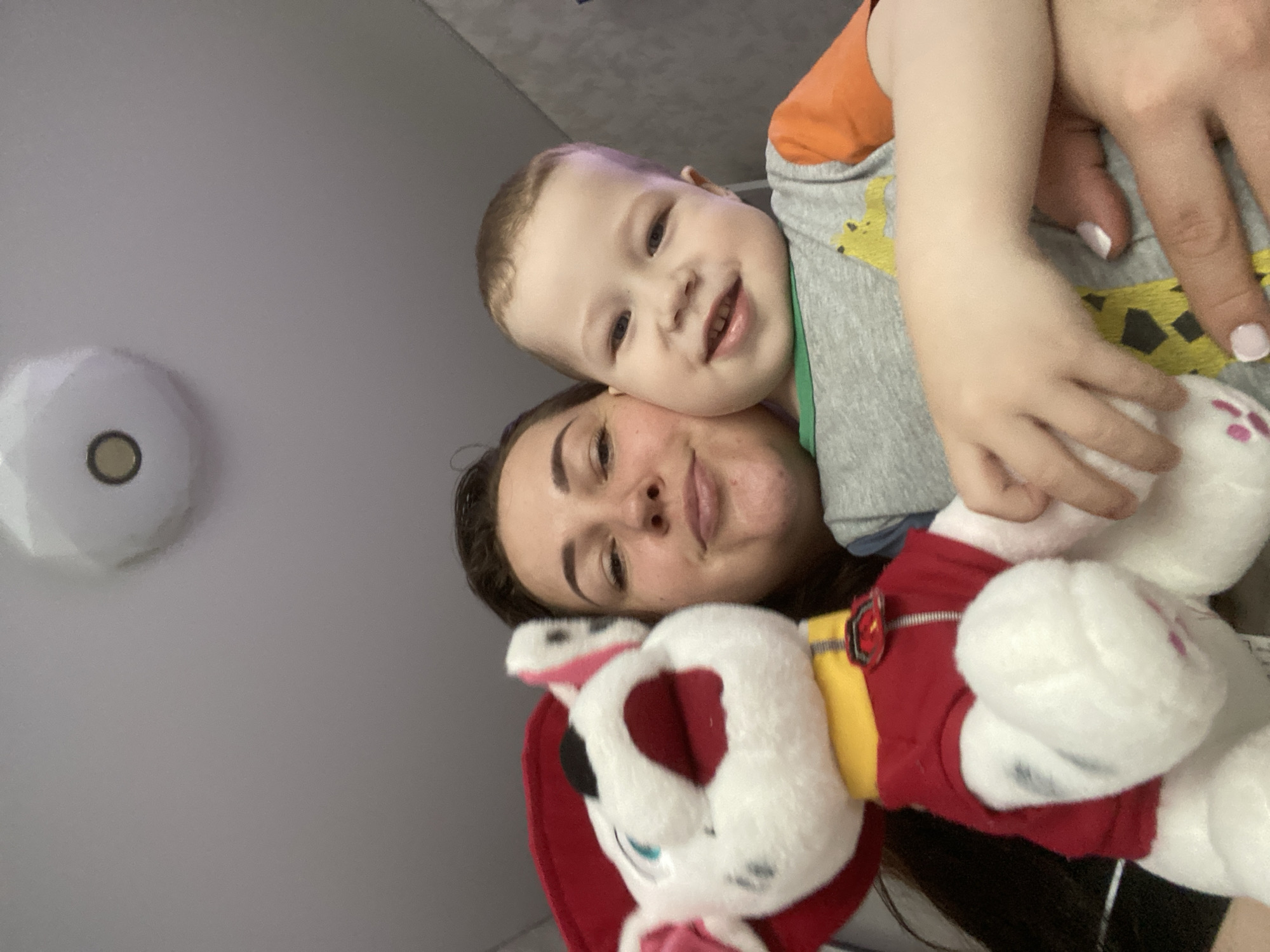 A woman and a young child are smiling at the camera. The child is wearing a grey shirt with an orange sleeve and a green collar. The woman is holding a plush toy of Marshall from Paw Patrol. A ceiling light fixture is visible in the background. The location appears to be indoors, possibly a home. The city is not clear.