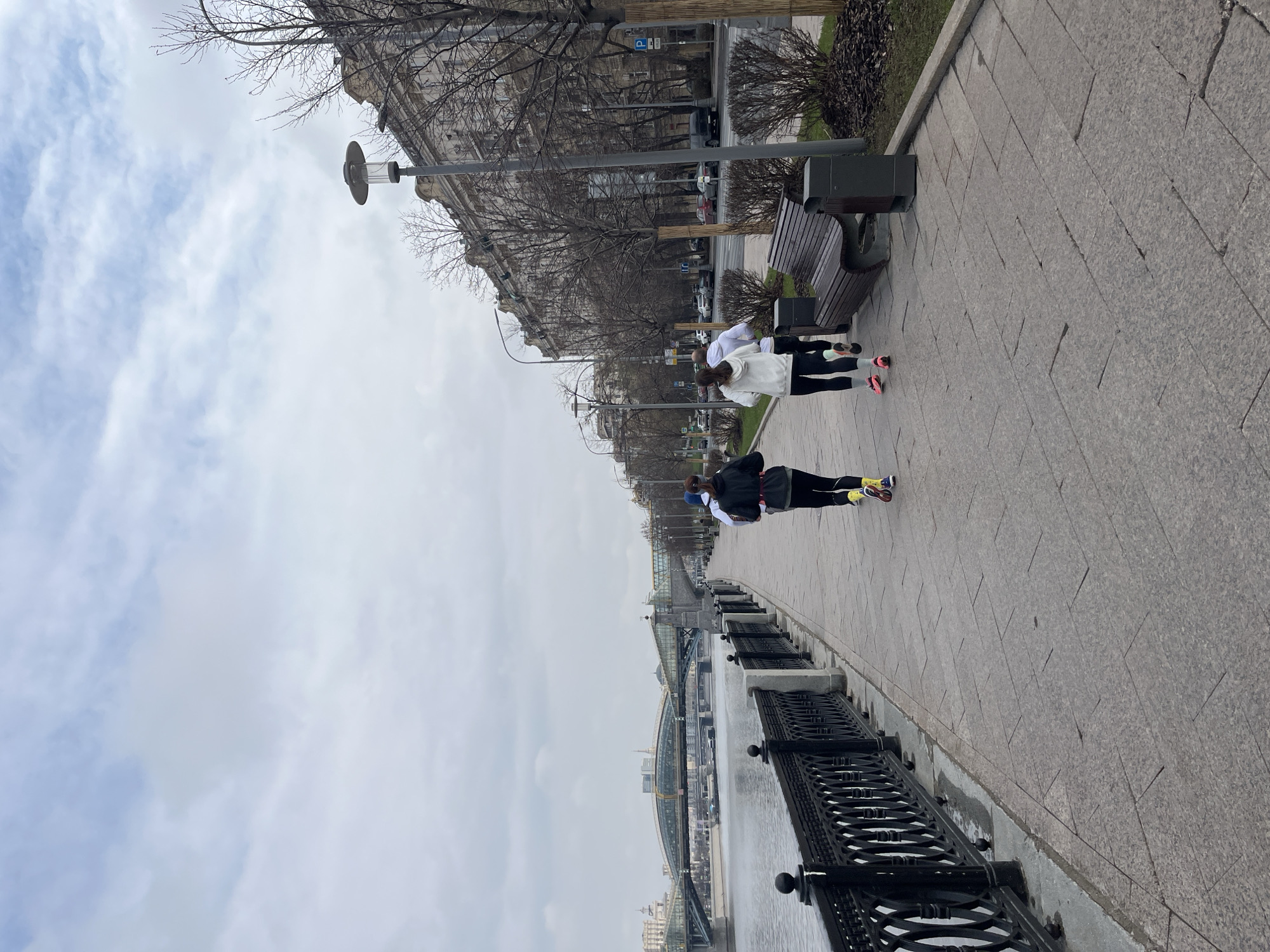 The image shows a Moscow, Russia riverside scene. Three people are walking along a paved sidewalk next to a river with a black iron railing. The person in the front is wearing a black vest, white shirt, black leggings, and yellow and blue sneakers. The other two people are wearing white hooded sweatshirts and black leggings with pink sneakers. To the left of the walkers is the river, where two bridges can be seen in the distance. To the right is a row of buildings, lamp posts, and a park bench. The sky is overcast.
