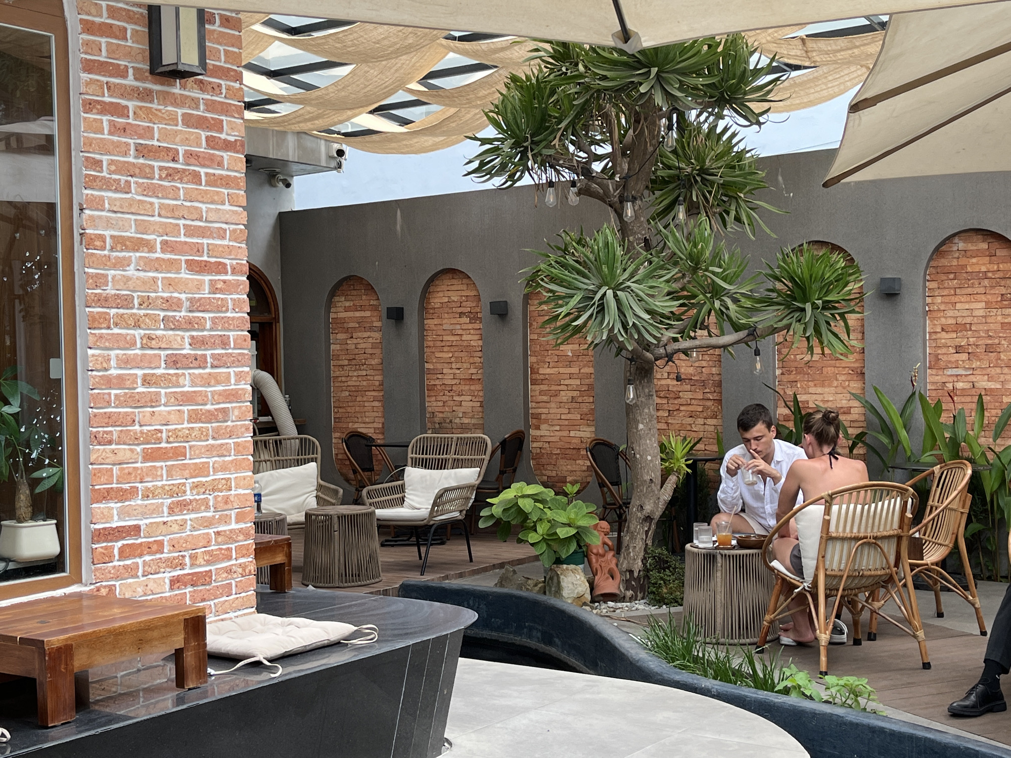 This image captures an inviting outdoor café or restaurant scene, characterized by a blend of rustic and modern design elements. The setting features exposed red brick walls and pillars contrasting with smooth dark grey walls, some of which incorporate arched alcoves, two filled with brick.
In the mid-ground, a man and a woman are seated at a low, round table made of wicker-like material, enjoying drinks. The man, wearing a white button-up shirt and light shorts, is looking down at his drink. The woman, seen from behind, wears a dark bikini top and shorts, and is seated in a matching wicker chair with a white cushion. They appear relaxed and engaged in conversation. Behind them, lush green foliage adds a natural touch.
A prominent feature in the center is a large, sturdy tree with long, spiky green leaves, from which string lights with bare bulbs are suspended. Below the tree, a small potted plant and a terracotta figurine are visible. Overhead, a beige fabric canopy provides shade, creating a dappled light effect, while a large white umbrella is also visible on the right.
Other elements include various wicker chairs and tables arranged around the space, some with white cushions. In the foreground on the left, a dark wooden bench sits beside a brick wall, with a white cushion resting on a dark surface, possibly part of a low wall or water feature that curves into the frame. The overall atmosphere is tranquil and comfortable, suggesting a pleasant daytime experience. No visible text is present.