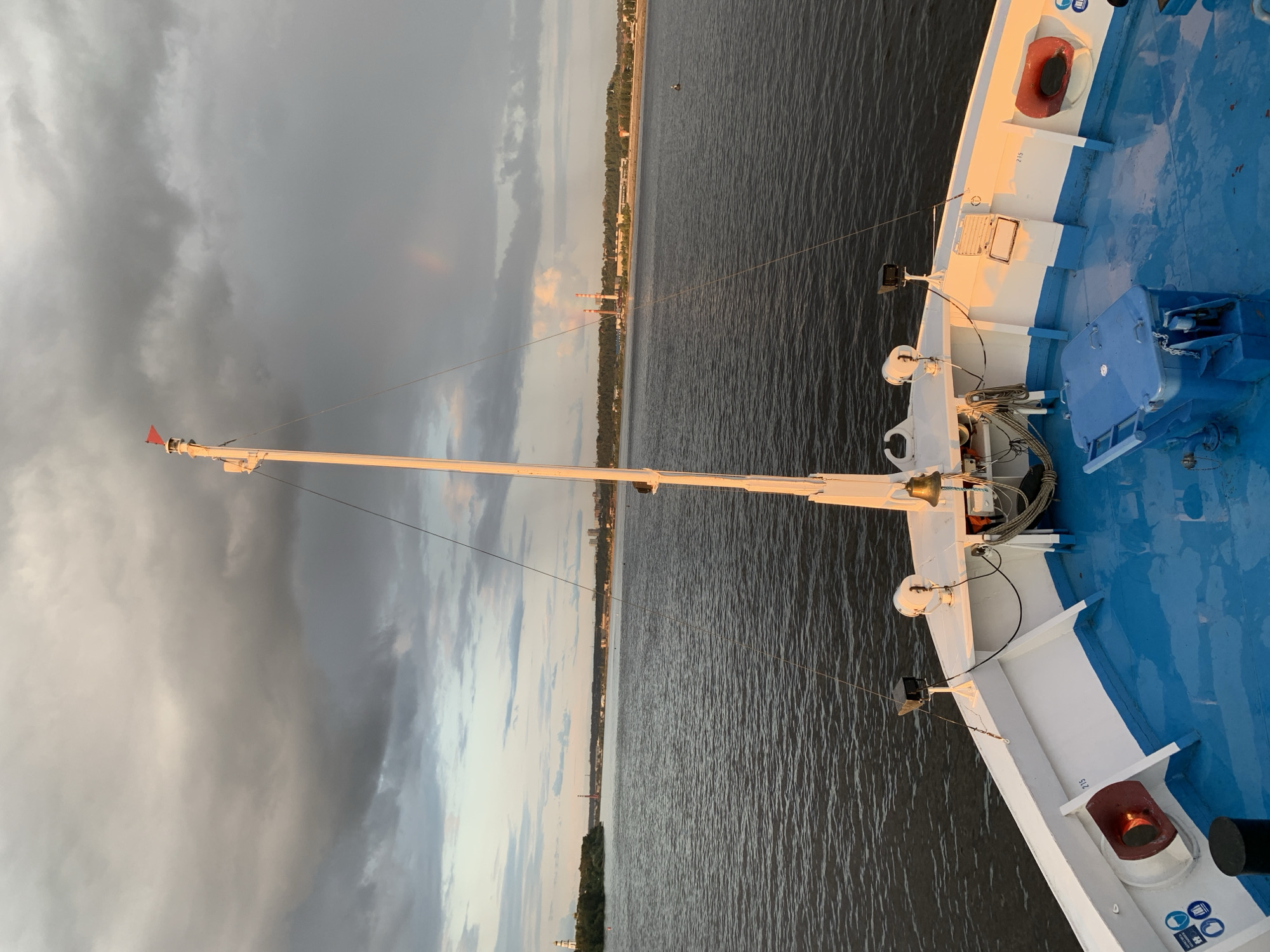 The image shows the bow of a white ferry with a bright blue deck, navigating a wide, dark river. A tall white mast with a red flag at its apex rises from the deck, with rigging lines extending outwards. A bell is mounted on the deck near the mast. In the background, a tree-lined shore is visible, with industrial buildings and chimneys spewing smoke, indicating a likely industrial area of the city of Perm, Russia. A lighthouse is visible on the left bank. The sky is dramatic, with heavy, dark clouds casting a somber mood, yet a faint rainbow is visible in the distance, suggesting recent or ongoing rain. The sunlight, low on the horizon, casts a warm glow on parts of the ship's deck and the distant industrial structures. The water is calm, reflecting the subdued lighting conditions. There are no people visible in the image.