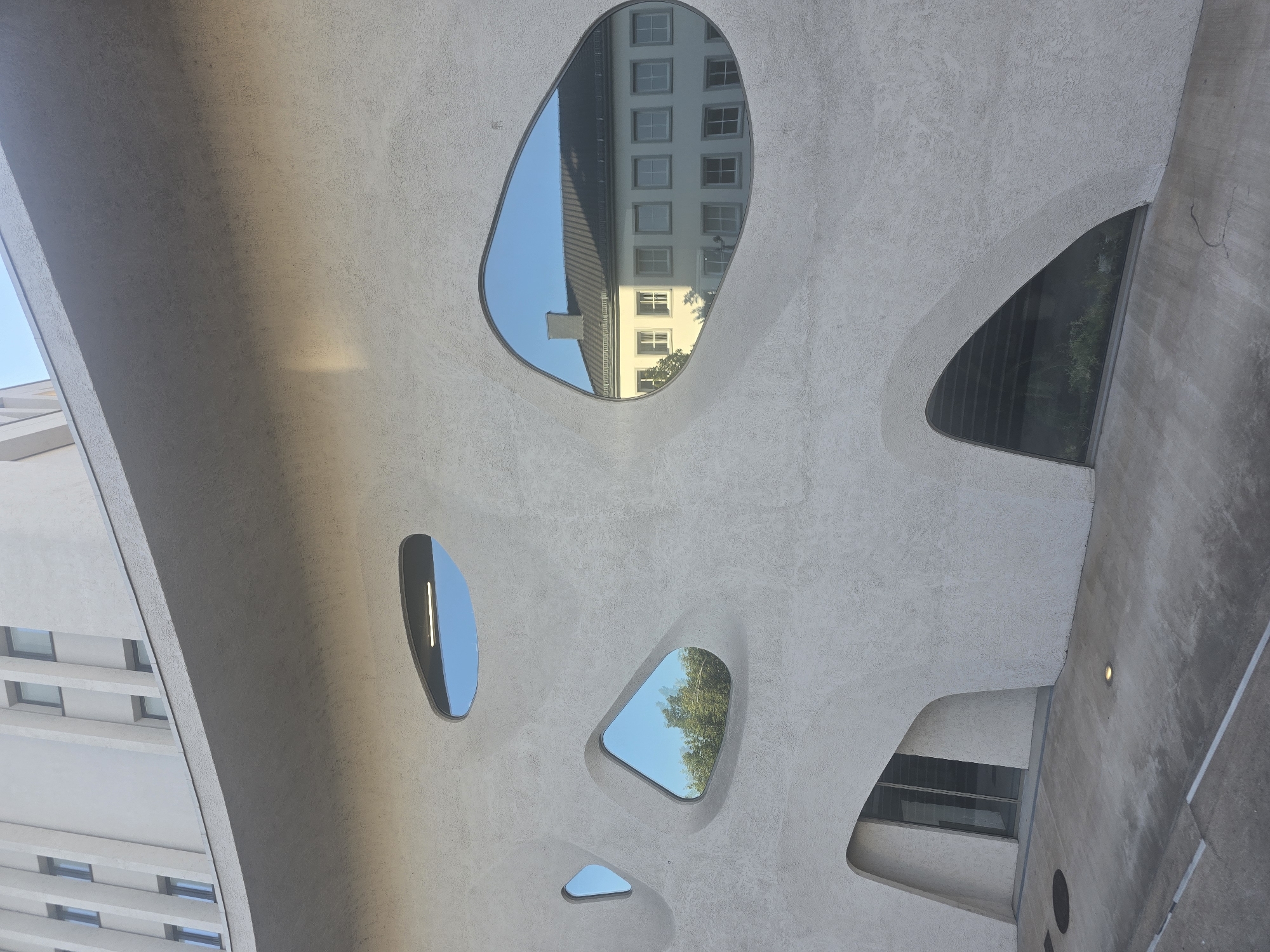 The image depicts the exterior of a modern building with an unusual, organic facade in Saarlouis, Germany. The building's exterior is textured and light gray, with several irregularly shaped windows that act as openings. Some windows are small and triangular, while others are larger and more elongated, resembling amoeba shapes.
One of the larger windows reveals a reflection of a pale yellow building with multiple windows and a dark gray roof. This reflected building appears to be traditional in architectural style, contrasting with the modern building in the foreground. Another window reflects a clear blue sky and the green foliage of trees.
At the bottom left of the building, there is an entrance with glass doors set within a curved opening. The ground in front of the building is a concrete walkway with some subtle markings. Several small, recessed lights are visible on the concrete surface. To the right, there is a large, triangular window at ground level that looks into an interior space with visible rows of stairs and some greenery.
The overall impression is one of contemporary architecture integrating with its surroundings through reflections and organic forms. The scene is well-lit, suggesting it was captured during daylight hours, likely in the morning or late afternoon due to the angle of the light and shadows. No people are visible in the image.