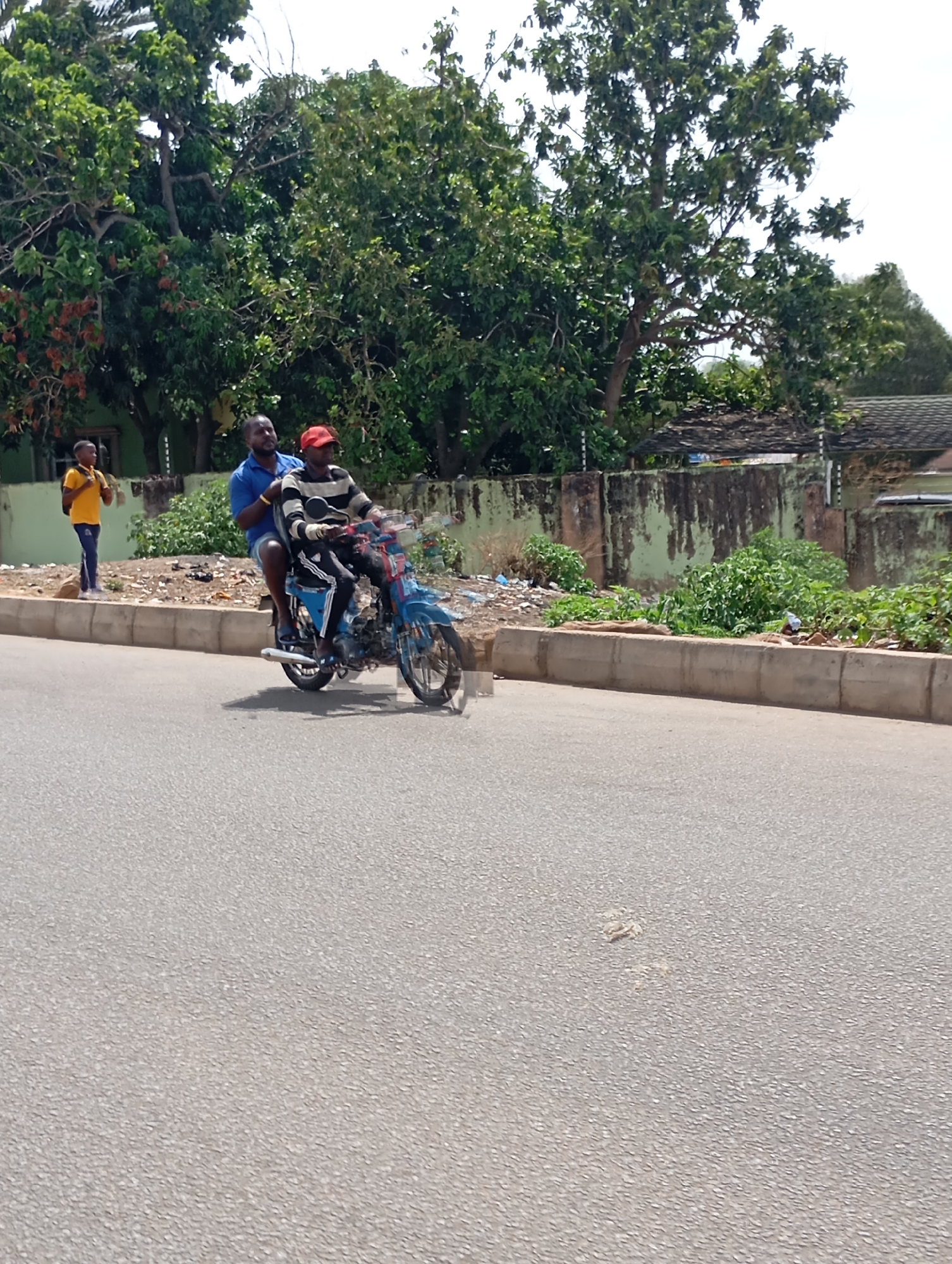 This daytime image, set in Jos, Nigeria, captures a street scene featuring two men on a blue motorcycle riding on an asphalt road. The driver wears a black and white striped long-sleeve shirt, dark pants with white stripes, and a red cap. A passenger sits behind him, dressed in a blue short-sleeve polo shirt and dark shorts. To the left, a child or young person in a yellow shirt and dark pants stands on the roadside, behind a concrete curb, observing the motorcycle.
The road is paved, and a concrete curb separates it from a rough verge containing scattered litter and sparse green vegetation. In the background, a low concrete wall, discolored by patches of moss or grime, stands before a dense line of lush green trees, some exhibiting reddish-brown foliage. The bright, possibly hazy sky suggests sunny weather conditions. The scene appears to be an ordinary moment of daily life, with no strong emotions evident. No text is visible within the image.