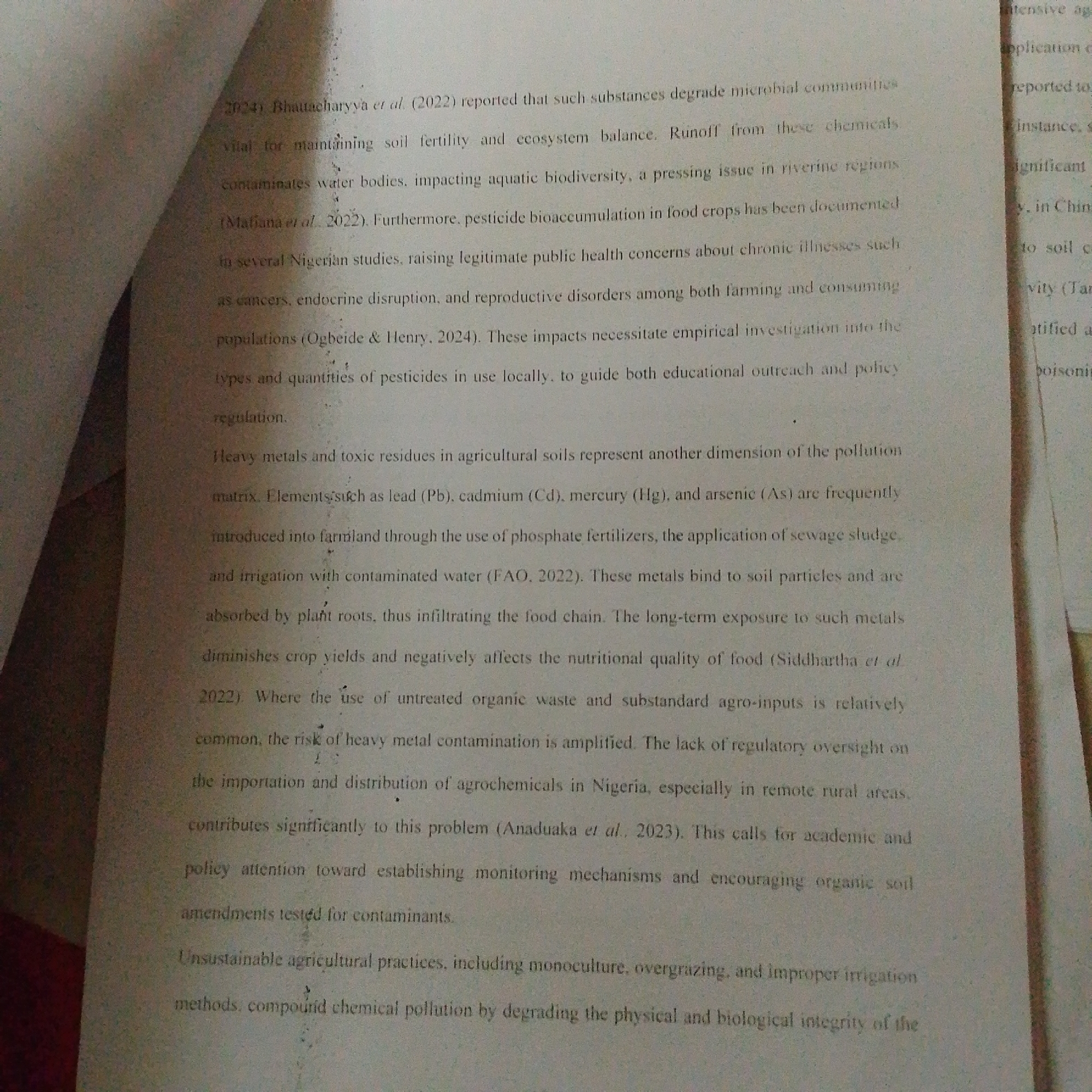 This media file is a close-up shot of a document page, likely from an academic paper or report. The text is printed in black on a light-colored paper, which appears slightly aged or creased, with a visible shadow or fold on the left side, obscuring some of the text.
The document discusses various aspects of agricultural pollution, specifically focusing on the impact of pesticides and heavy metals. It references research and studies, citing authors and publication years such as Bhattacharyya et al. (2022), Mafiana et al. (2022), Ogbeide & Henry (2024), FAO (2022), Siddhartha et al. (2022), and Anaduaka et al. (2023). The text details how these substances degrade soil fertility, contaminate water bodies, and bioaccumulate in food crops, leading to public health concerns like cancers and endocrine disruption. It also touches upon the role of phosphate fertilizers, sewage sludge, and contaminated irrigation water in introducing heavy metals into agricultural soils, and the subsequent impact on crop yields and nutritional quality. The document highlights the amplified risk of heavy metal contamination in Nigeria due to the importation and distribution of agrochemicals and the lack of regulatory oversight, especially in remote rural areas. It calls for academic and policy attention towards establishing monitoring mechanisms and encouraging organic soil amendments. Finally, it mentions unsustainable agricultural practices like monoculture, overgrazing, and improper irrigation methods as compounding chemical pollution.
The specific location mentioned in the context is Jalingo, Nigeria, suggesting the research or data discussed pertains to this region or has implications for it. No people, objects, or specific scenes are depicted other than the printed text itself. The time of day, weather, and emotions are not discernible from the content.