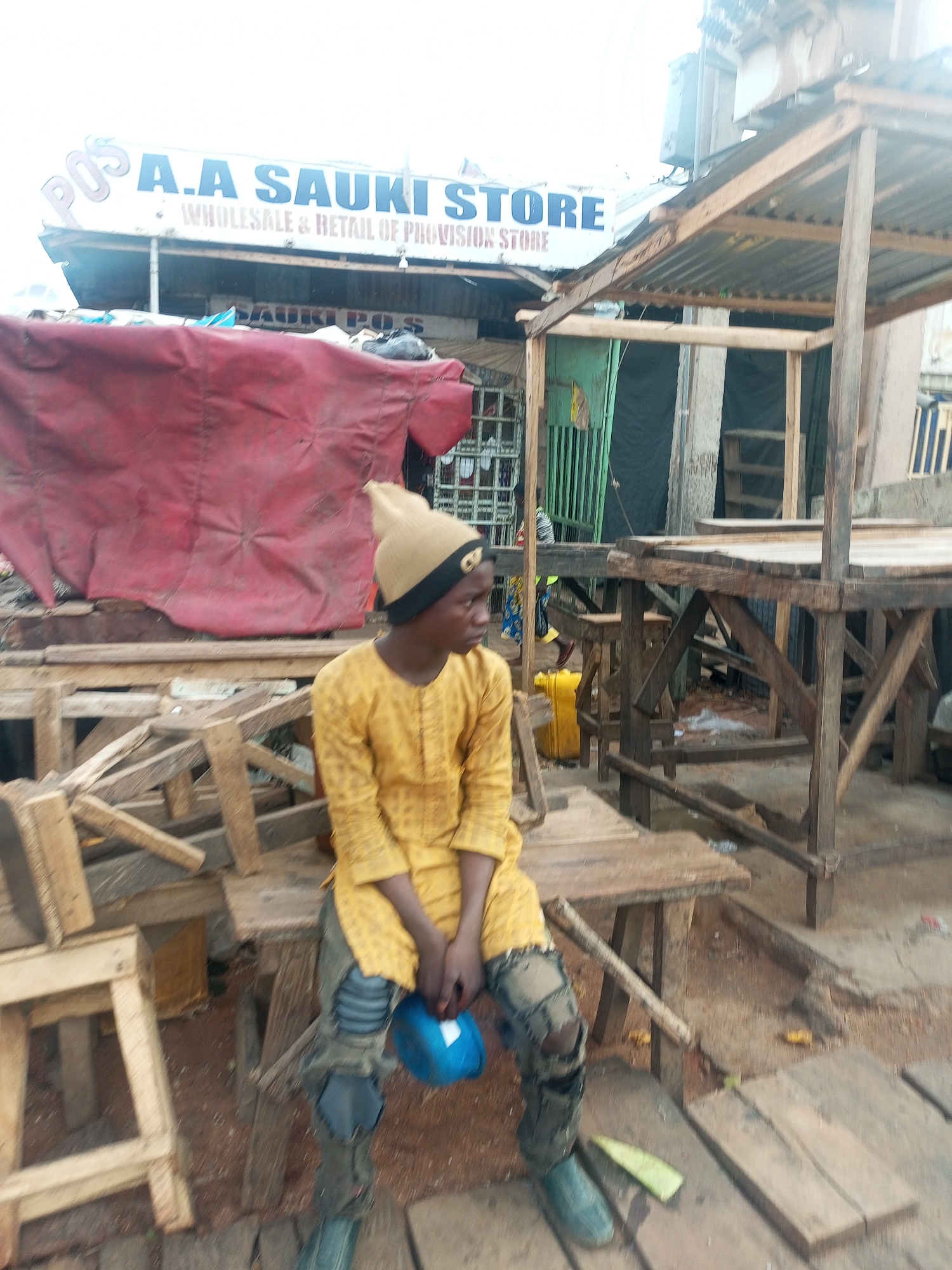 A young person sits on a rough wooden bench, holding a blue object. The person wears a tan hat, a yellow patterned shirt, and ripped jeans, with what appear to be patched-up boots. The background includes a store sign reading "A.A SAUKI STORE WHOLESALE & RETAIL OF PROVISION STORE," suggesting a commercial area. Red cloth is hanging over what appears to be a dilapidated market stall. Numerous roughly constructed wooden structures are visible, suggesting a market or construction site. The location is Tempure, Nigeria.