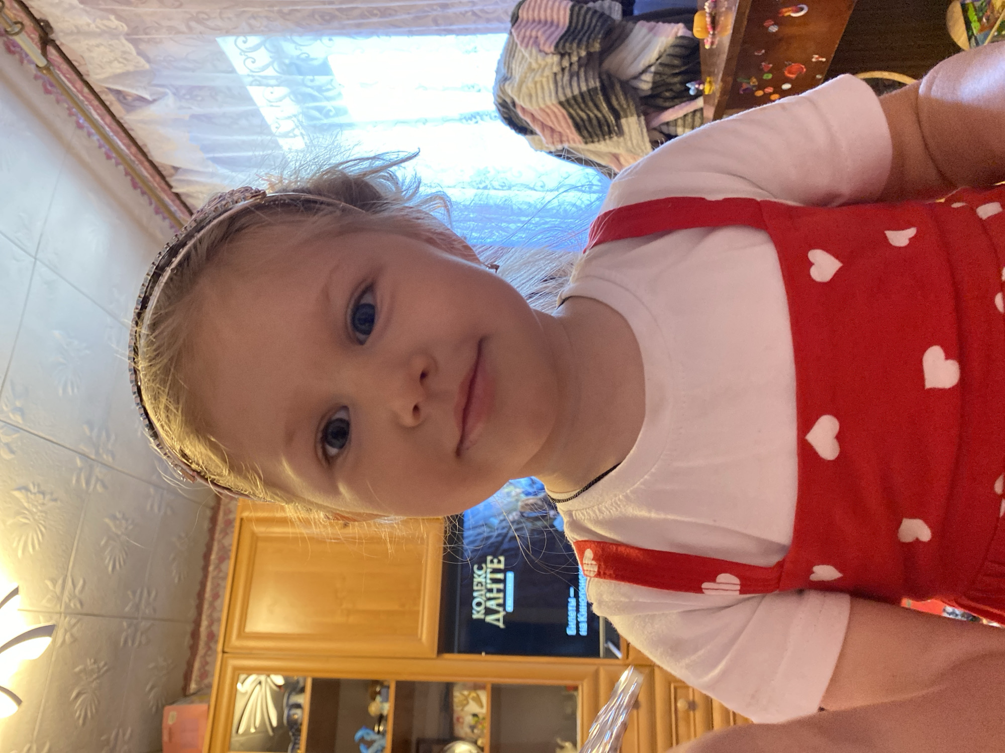 A young girl with blonde hair and blue eyes, wearing a white t-shirt and a red dress with white hearts, is pictured from the chest up. She has a headband with rhinestones in her hair. The background includes a wooden cabinet, a television displaying text in Russian that reads "КОДЕКС ДАНТЕ" (Dante's Code), and a window with lace curtains. A striped blanket is draped over furniture to the right. The scene appears to be indoors, possibly a living room. The location is identified as Mariupol, Ukraine.