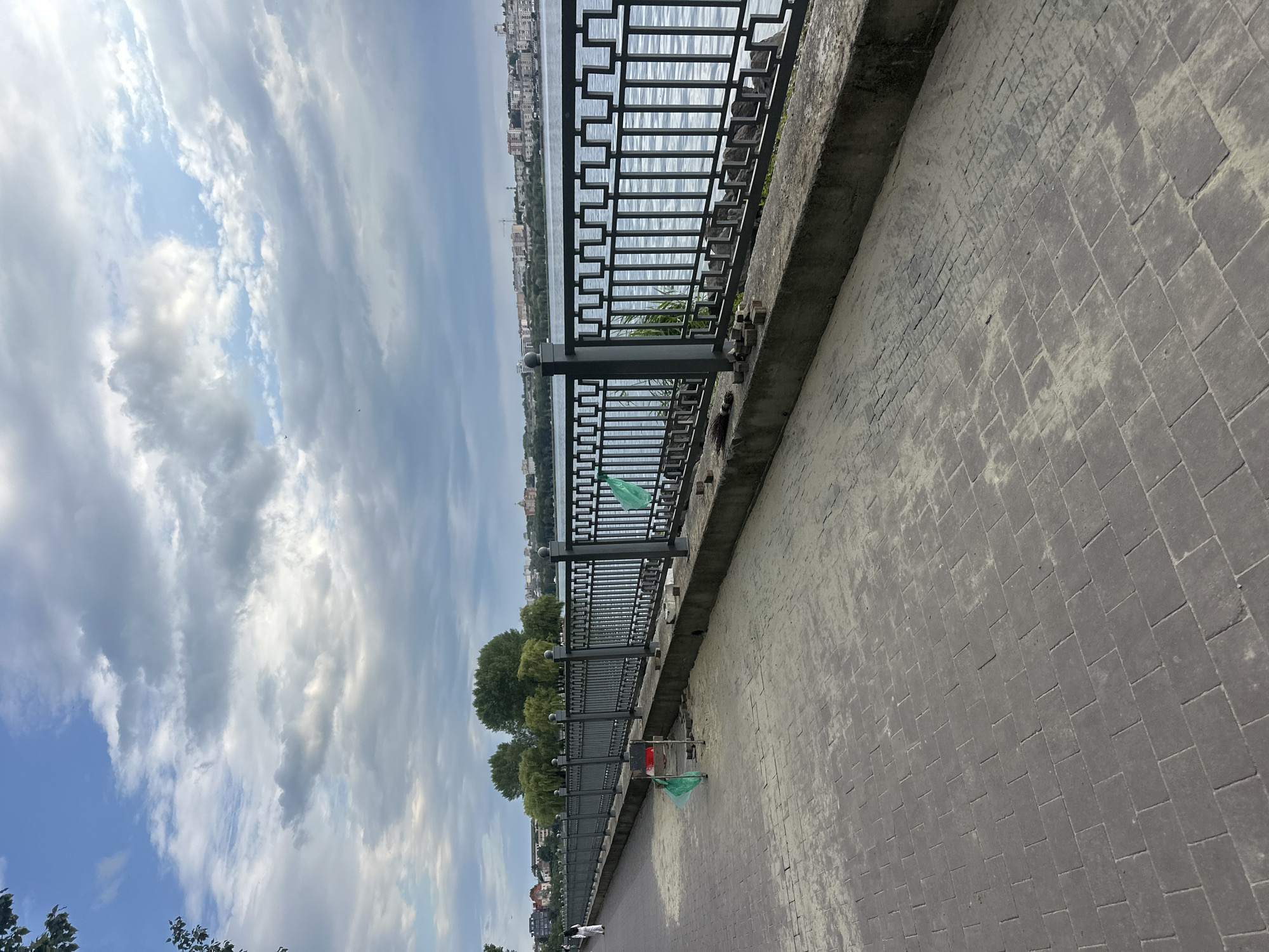 The image depicts a paved walkway adjacent to a body of water, likely Ternopil Lake, in Ternopil, Ukraine. A decorative metal fence separates the walkway from the water. The walkway is constructed of interlocking paving stones and appears to be undergoing some repair work, with piles of bricks and sand visible near the fence. Two small green flags on stands are placed on the walkway, possibly marking an area of work. In the distance, a city skyline with buildings and trees can be seen across the water. The sky is partly cloudy. One person is visible walking on the far side of the walkway. The scene captures a public space along the waterfront.