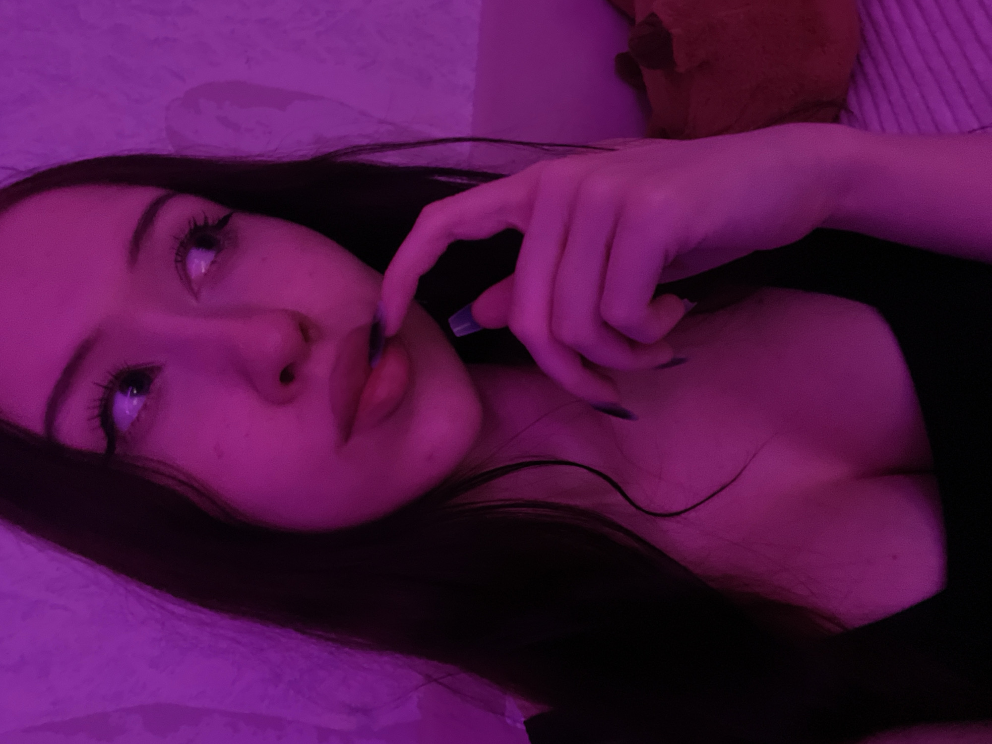 A young woman with dark hair and makeup is captured in a close-up shot, illuminated by vibrant pink lighting. Her left eye is visible, looking to the side, and her lips are slightly parted as she brings her right hand to her mouth. She is wearing a black top that reveals a portion of her chest. The background appears to be a textured wall, also bathed in the pink light. There is no discernible information within the image that places it in Votkinsk, Russia.