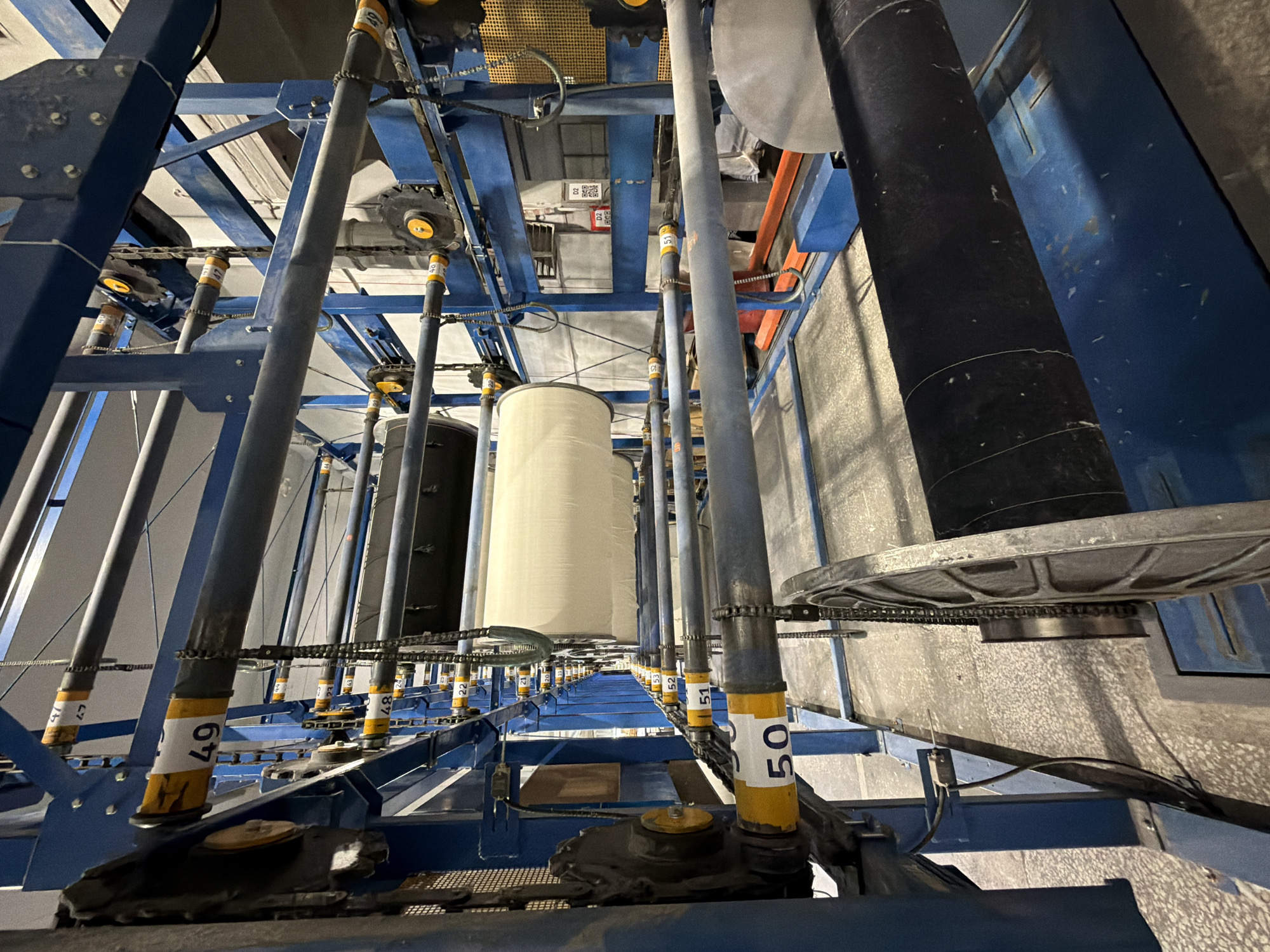 The image shows an industrial setting in what could possibly be a production or storage facility.
**Objects**: There are rows of vertical cylindrical objects that appear to be large spools or rolls, some white and others black. These objects are supported by a complex blue metal frame structure with chains and numbered yellow and white labels (e.g., "50", "49"). The lighting is artificial and comes from above, casting shadows. There are also gray metal panels with the spools attached to them.
**Environment**: The scene appears to be inside a tall industrial building.
**Location and time**: There is nothing to verify the location as Chaikovsky, Russia. It is inside so the time of day can not be determined.