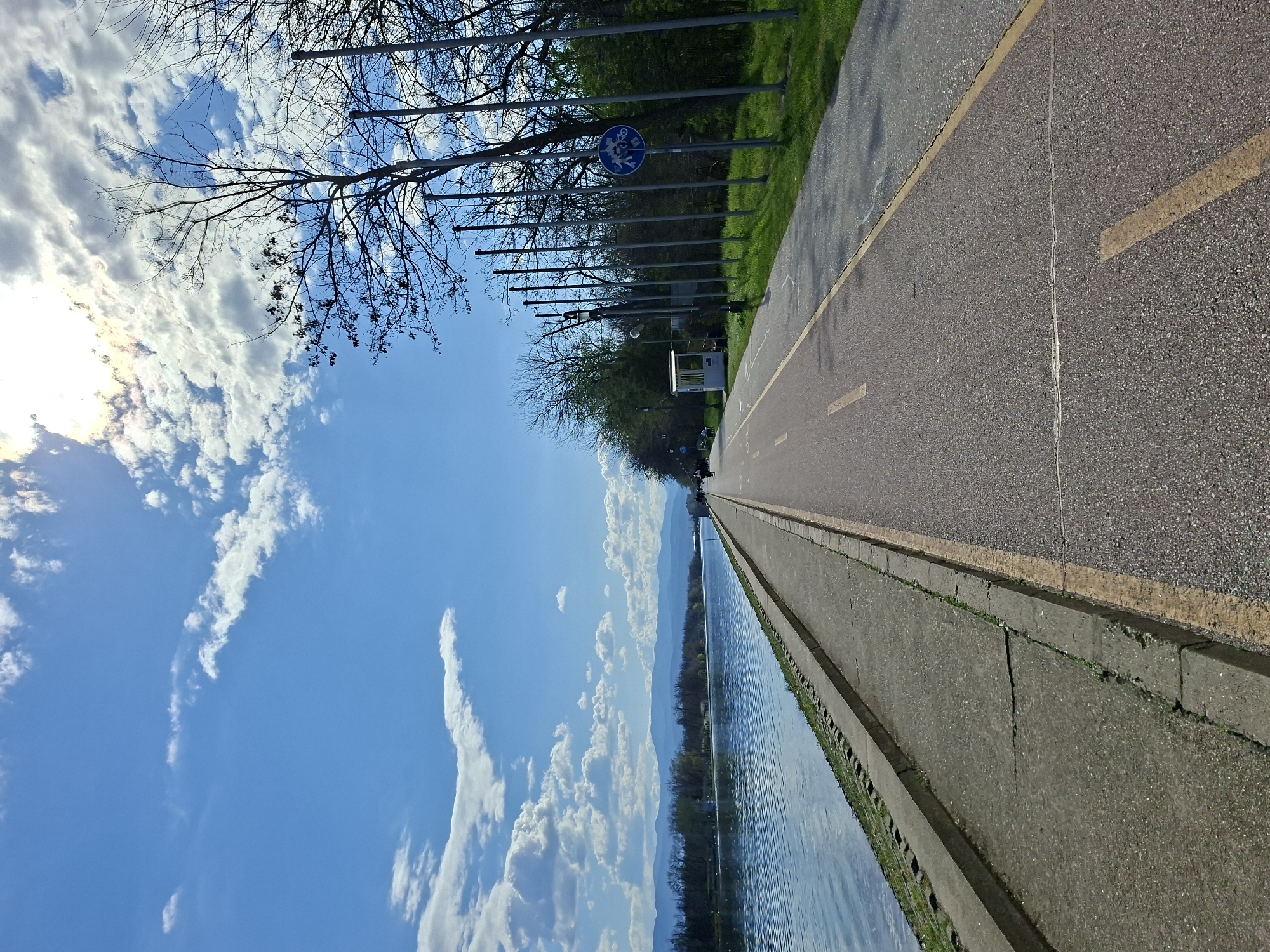 The image depicts a paved path in Plovdiv, Bulgaria, adjacent to a body of water. The path has a yellow line marking. To the left of the path, there's a concrete barrier separating it from the water. On the opposite side of the path, a row of metal posts runs along a grassy area. Bare trees and some with budding leaves are visible near the posts. In the background, distant mountains are seen across the water. The sky is blue with scattered white clouds, and the sun is visible, creating a bright spot in the upper left. A blue circular sign with bicycle symbols is mounted on one of the metal posts. No people are clearly visible on the path.