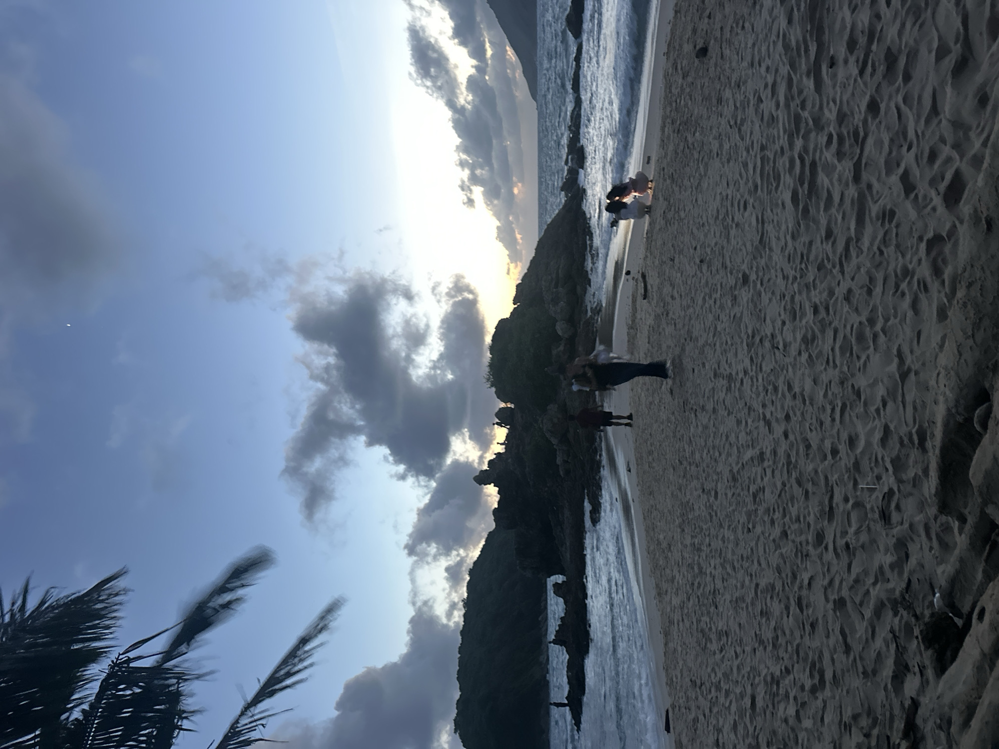 A group of people are on a sandy beach in Rawai, Thailand, near a rocky outcrop in the water. The sun is setting, casting a warm glow on the clouds and the ocean. A palm tree is visible in the foreground, and the waves are crashing on the shore. Some individuals are walking along the beach, while others are sitting and observing the sunset. The overall atmosphere is peaceful and serene.