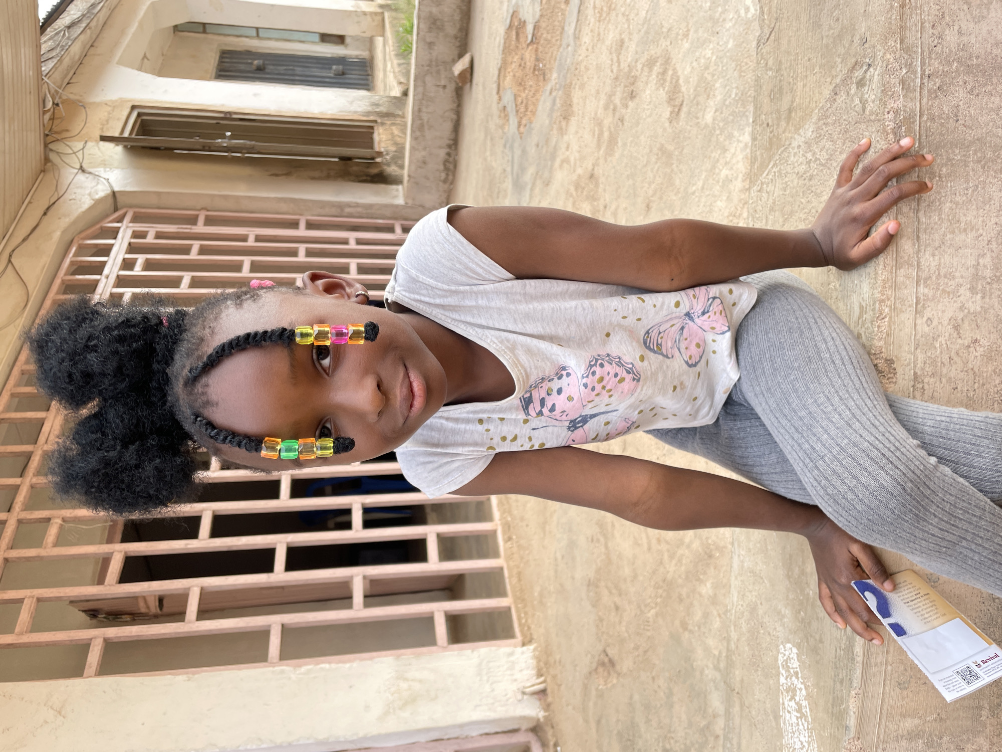 A young Black girl is captured in a close-up, three-quarter length shot, looking directly at the camera with a gentle expression. She is seated outdoors on a light-colored paved or tiled surface, against the backdrop of a building. Her dark, curly hair is styled in two puffs on top of her head, with small braids framing her face. These braids are adorned with colorful, square, translucent beads in shades of orange, green, yellow, and pink. She wears a white t-shirt decorated with pink and gold butterfly motifs, paired with gray ribbed leggings. Her right hand rests flat on the ground beside her, while her left hand holds a small, folded brochure or pamphlet.
The setting appears to be an outdoor area in Barakin Akawo, Nigeria, characterized by worn, light-colored building walls and a robust, light-colored barred window or gate in the background. The bright, natural light indicates it is daytime, casting soft shadows. The object in her hand clearly shows the word "Revival" and a QR code, alongside other smaller, illegible text. The overall scene depicts a casual, everyday moment.