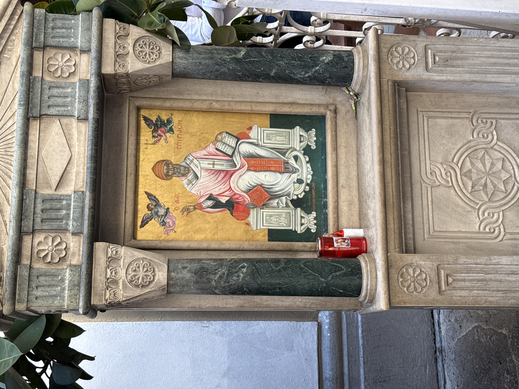 The image shows an outdoor religious shrine in the city of Corfu, Greece. The shrine is constructed from stone and features an icon of Saint Spyridon, the patron saint of Corfu. The icon depicts Saint Spyridon seated and holding a book, with two angels on either side. To the left of the icon, there is a red votive candle with a depiction of the Virgin Mary and child. There are also two small flowers placed on the ledge. The shrine is partially obscured by green foliage from a nearby tree. The background includes a light blue wall and a white ornate metal railing. The shrine is positioned on a stone pedestal with decorative carvings.