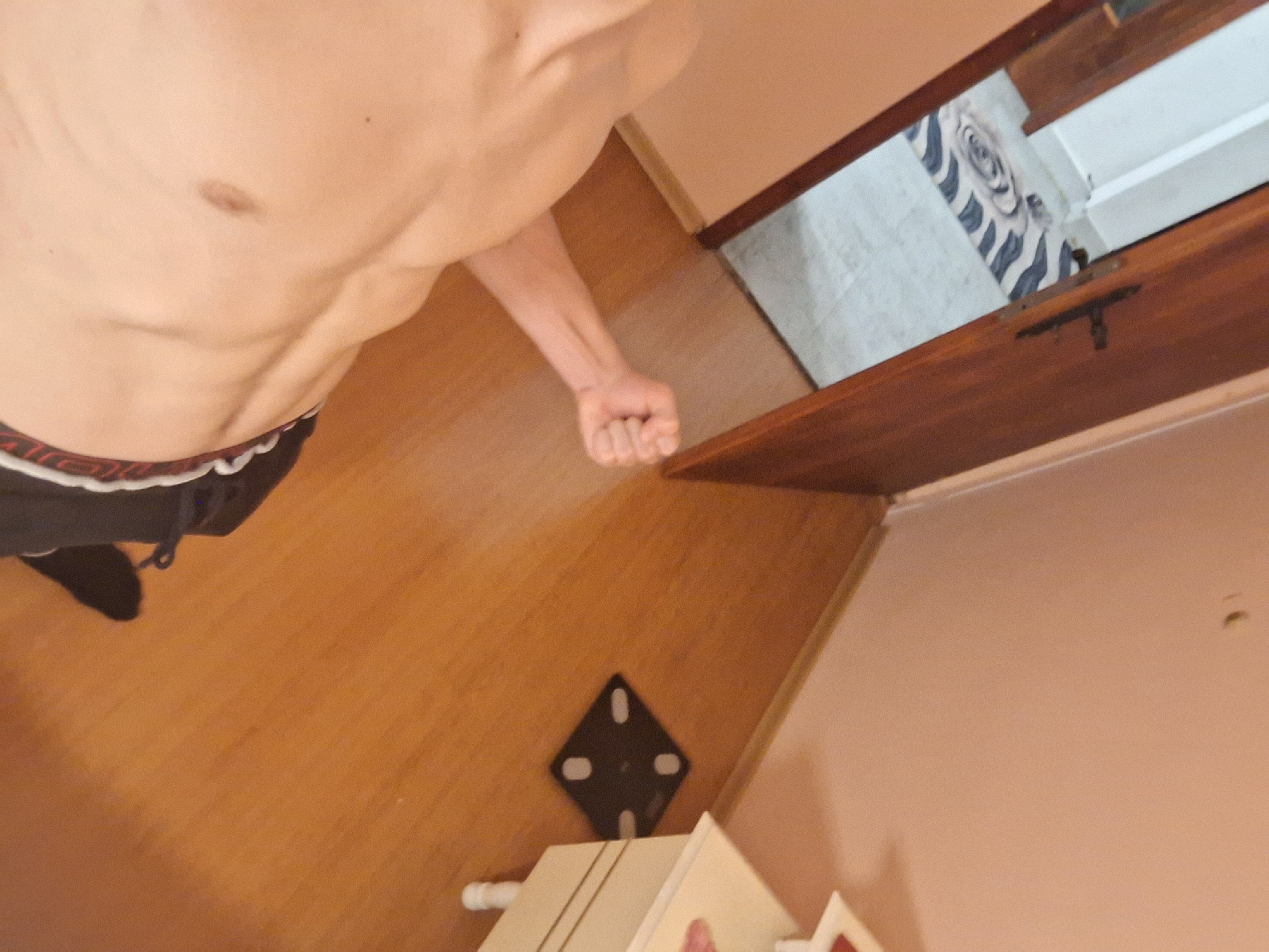 A shirtless male with visible abdominal muscles is standing in a room, potentially a bedroom or living space, as indicated by the hardwood flooring. He is wearing black bottoms with a red and white waistband and black socks. His right arm is extended with a clenched fist.
In the background, there is a partial view of a room with a rug featuring a patterned design of gray and white. A wooden door is slightly ajar.
On the floor, there is a small black device with four white dots, possibly a digital scale or a similar electronic device. In the lower part of the image, there is a portion of what appears to be a white piece of furniture.
The image is taken from a high angle, looking down, and the lighting seems to be artificial. The location in Burgas, Bulgaria, cannot be determined from the visual information alone.