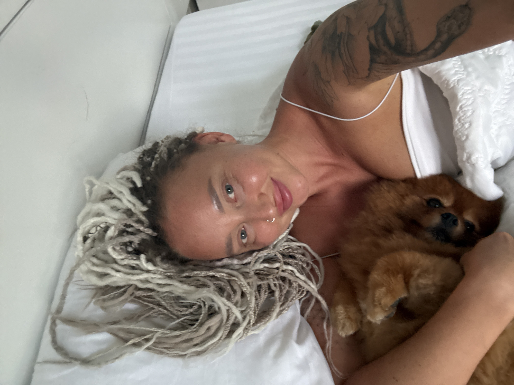 The media file shows a close-up, overhead shot of a person lying in bed with a small, light brown dog. The person has light-colored dreadlocks and a tattoo on their arm. They are wearing a white top, and a thin white string is visible around their neck. The person is looking at the camera with a slight smile. The dog, a Pomeranian breed, is nestled on the person's chest. The setting appears to be a bedroom, with white, striped bedding. The lighting suggests it could be daytime. The overall mood is relaxed and intimate. No visible text is present in the image. The context provided places this scene in Bac Nha Trang Ward, Vietnam.