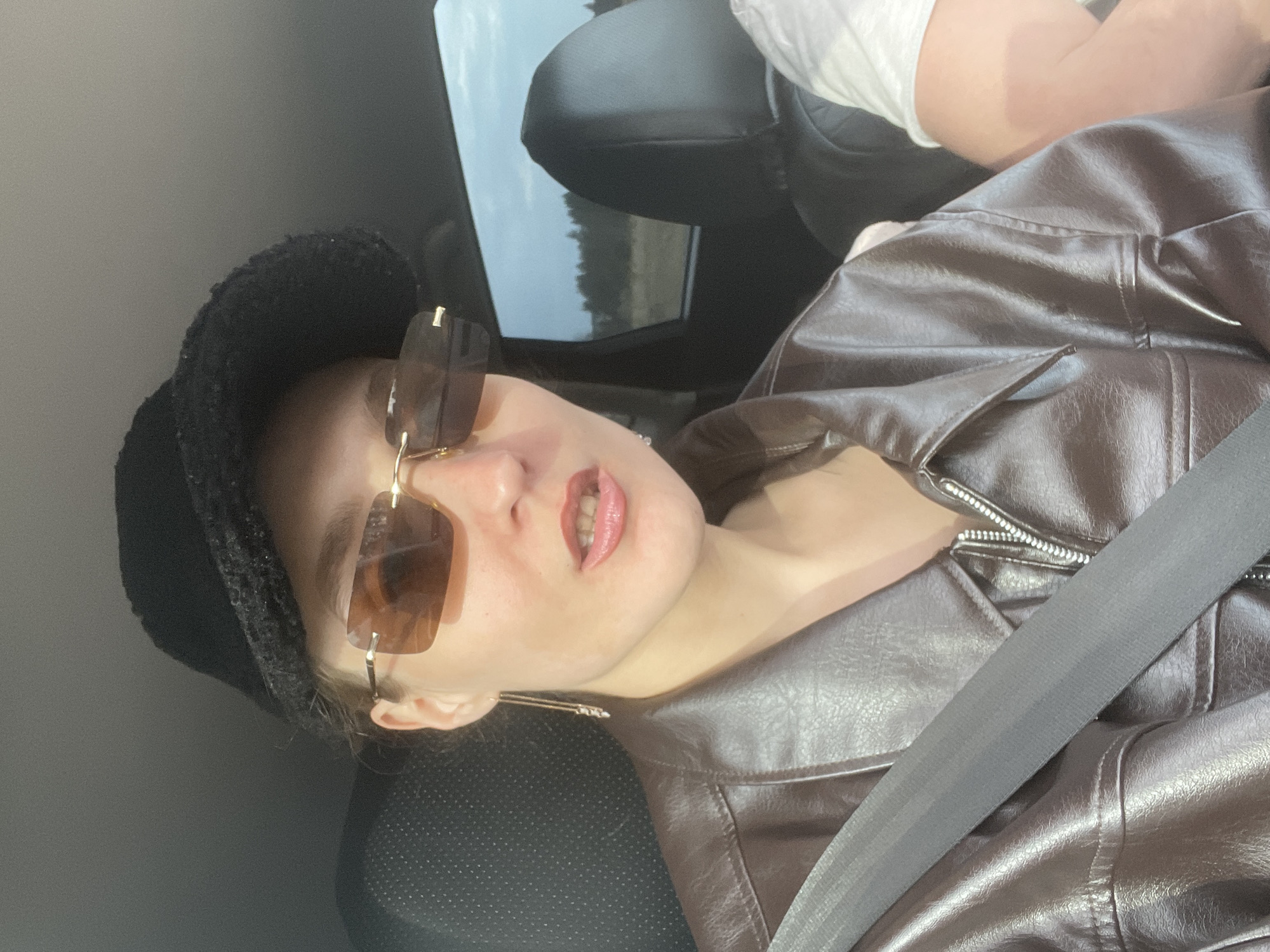 A person is seated in the front seat of a car, facing the camera. The image is taken indoors within a vehicle, identifiable by the dark car interior, seats, and a prominent dark gray seatbelt crossing the subject's chest and shoulder.
The individual, likely female, wears a dark, possibly black, fuzzy-textured hat. Their eyes are obscured by large, rimless, rectangular sunglasses featuring brown-tinted lenses and delicate gold-colored metal frames. They are dressed in a brown faux leather jacket, detailed with visible stitching and a silver-toned zipper on the front. Thin, gold-colored dangling earrings are visible below the hat.
The person's lips are painted with a reddish-pink lipstick and are slightly parted, revealing a glimpse of teeth. In the upper right corner of the frame, the arm of another individual, clad in a white short-sleeved top, is partially visible, suggesting the presence of another occupant in the car. Through the car window on the left, a blurred daytime landscape, likely comprising trees or natural scenery under a bright sky, can be seen. The overall lighting is bright, casting reflections on the sunglasses and jacket. The subject maintains a neutral expression while posing for the photograph. There is no visible text in the image.