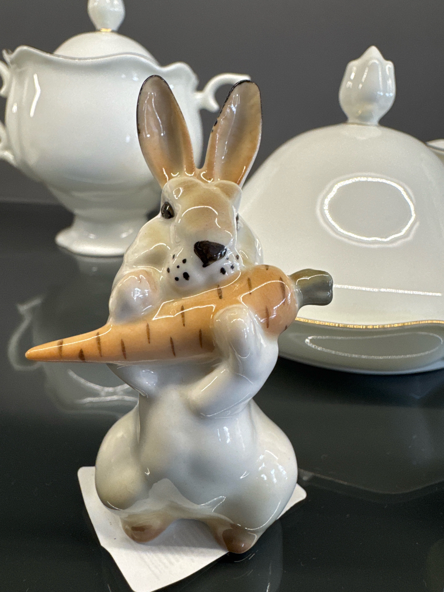 A close-up, eye-level, outdoor shot shows a ceramic figurine of a light brown and white rabbit holding an orange carrot in its paws. The rabbit figurine is detailed, with black painted dots on its cheeks, black eyes, and a black nose. Its ears are long and pointed, with a darker brown interior. The carrot is depicted with orange segments and green tops. The figurine is placed on a white paper tag, which is on a dark, reflective surface, possibly a table. In the background, out of focus, are two white ceramic objects with lids. The object on the left appears to be a teapot or sugar bowl with a scalloped edge and a decorative lid. The object on the right is a round lid with a gold rim and a floral-shaped finial. The background is a plain, dark gray wall. The overall lighting is soft and diffused. The image is a still life composition. There are no people or animals present. This image was taken in Novosibirsk, Russia.