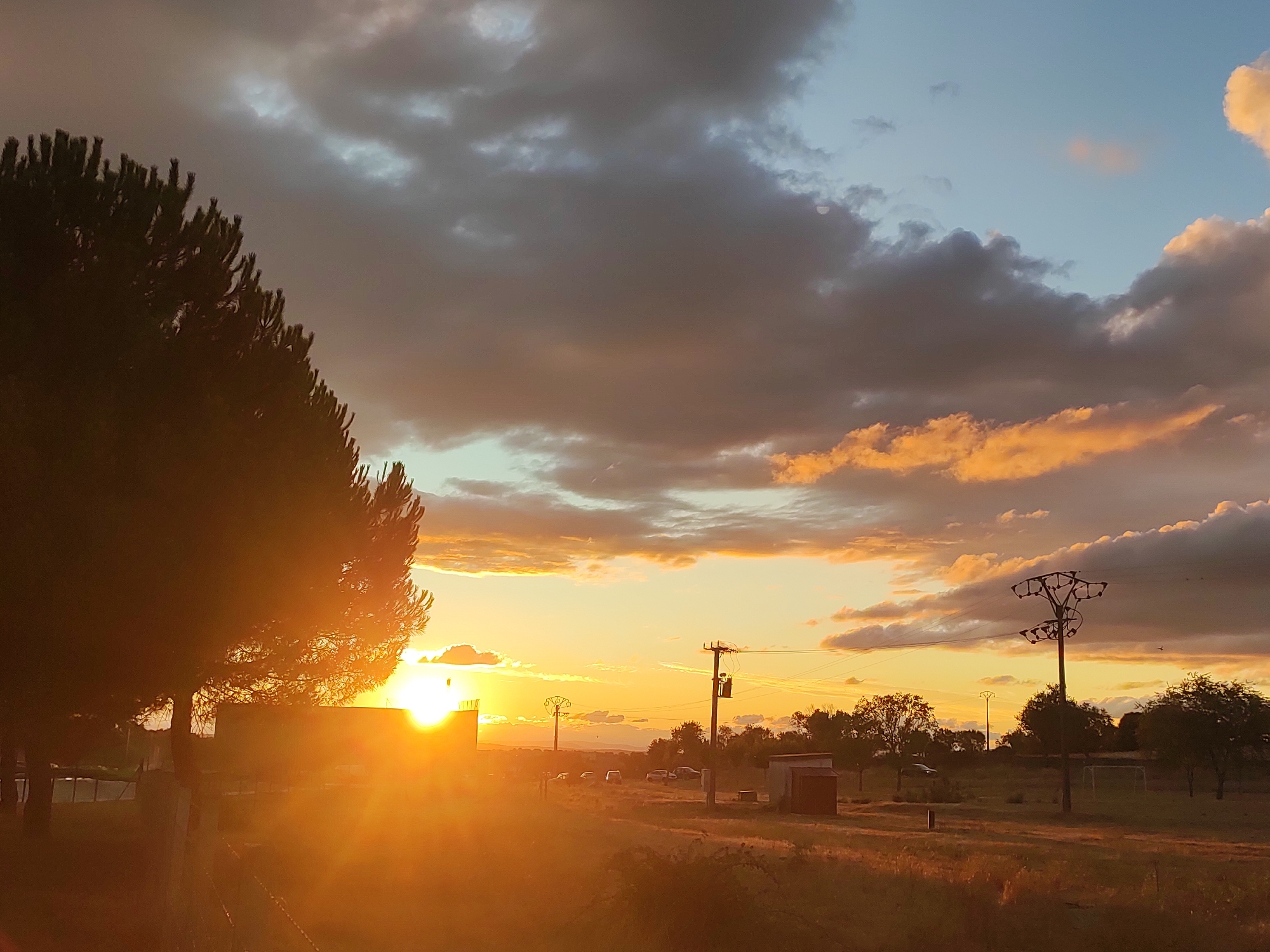 This image captures a dramatic sunset in the municipality of Sobradillo, Spain. The sun, a bright, intense disc, is setting behind a large, dark-silhouetted tree in the left foreground. The tree's foliage is rendered almost black against the vibrant orange and yellow hues of the sky.
The sky is a tapestry of contrasting colors, with deep grey storm clouds dominating the upper portion, interspersed with streaks of fiery orange and soft yellow where the sunlight breaks through. Towards the right of the frame, patches of clear blue sky are visible.
In the middle ground, there are several man-made structures. A low, rectangular building with a reddish-brown roof is visible on the right. Power lines and utility poles, some with transformers, traverse the landscape. A few vehicles, appearing as small shapes, are parked in the distance. A faint outline of a soccer goal can be seen in the far right.
The foreground consists of a field of dry, tall grass, rendered in warm, golden tones by the setting sun. A fence line is partially visible in the lower left corner. The overall impression is one of a rural landscape at dusk, with the natural beauty of the sunset juxtaposed against utilitarian infrastructure. The setting appears to be outdoors, likely in a semi-arid or rural area. There are no people visible in the image.