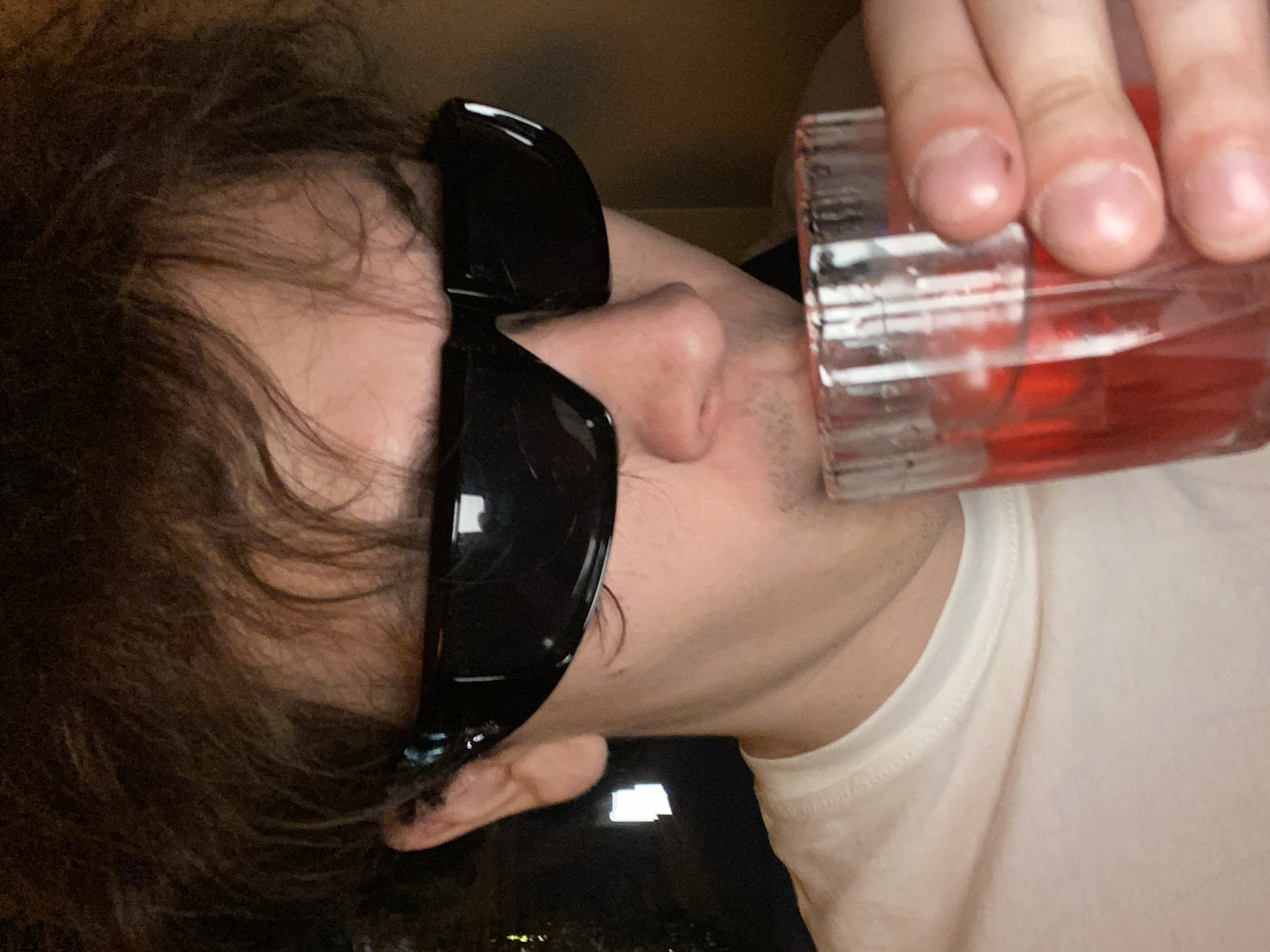 A person with dark, wavy hair and wearing dark sunglasses is shown in a close-up, angled shot. The person is drinking from a clear glass containing a red liquid. The person is wearing a white t-shirt. The background is blurry and dark. There is no discernible information to determine the location as Krasnoyarsk, Russia.