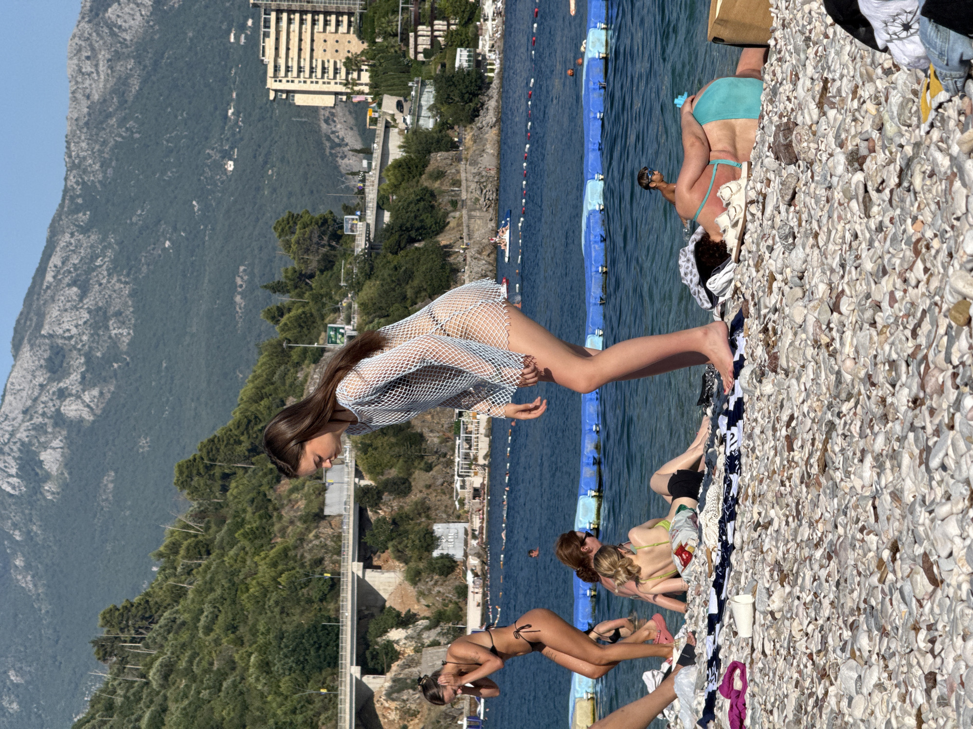 This media file shows a sunny day at a pebble beach in Budva, Montenegro. Several people are present, enjoying the beach and the sea. In the foreground, a young woman wearing a white mesh cover-up over a bikini is posing on the pebbles. To her right, another woman in a turquoise bikini is also on the beach, and further in the background, more people can be seen near the water, some swimming or relaxing on inflatables.
The scene is set against a backdrop of lush green mountains and a modern building complex, suggesting a coastal town. The water is a clear blue, and the sky is bright, indicating fair weather and likely midday. The overall atmosphere appears to be relaxed and recreational, with individuals enjoying a summer day by the sea. There are some beach accessories scattered around, like towels and a white bucket. No visible text is discernible in the image.