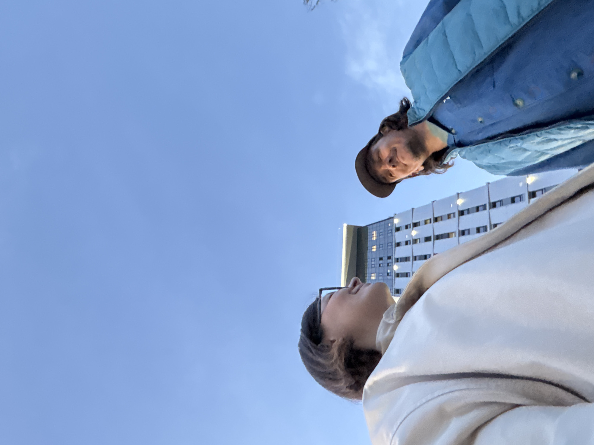 Two people are pictured from a low angle against a blue sky and a building. One person wears a blue jacket and a brown hat. The other person wears glasses and a white coat. The location appears to be Izhevsk, Russia, based on the architectural style of the building.