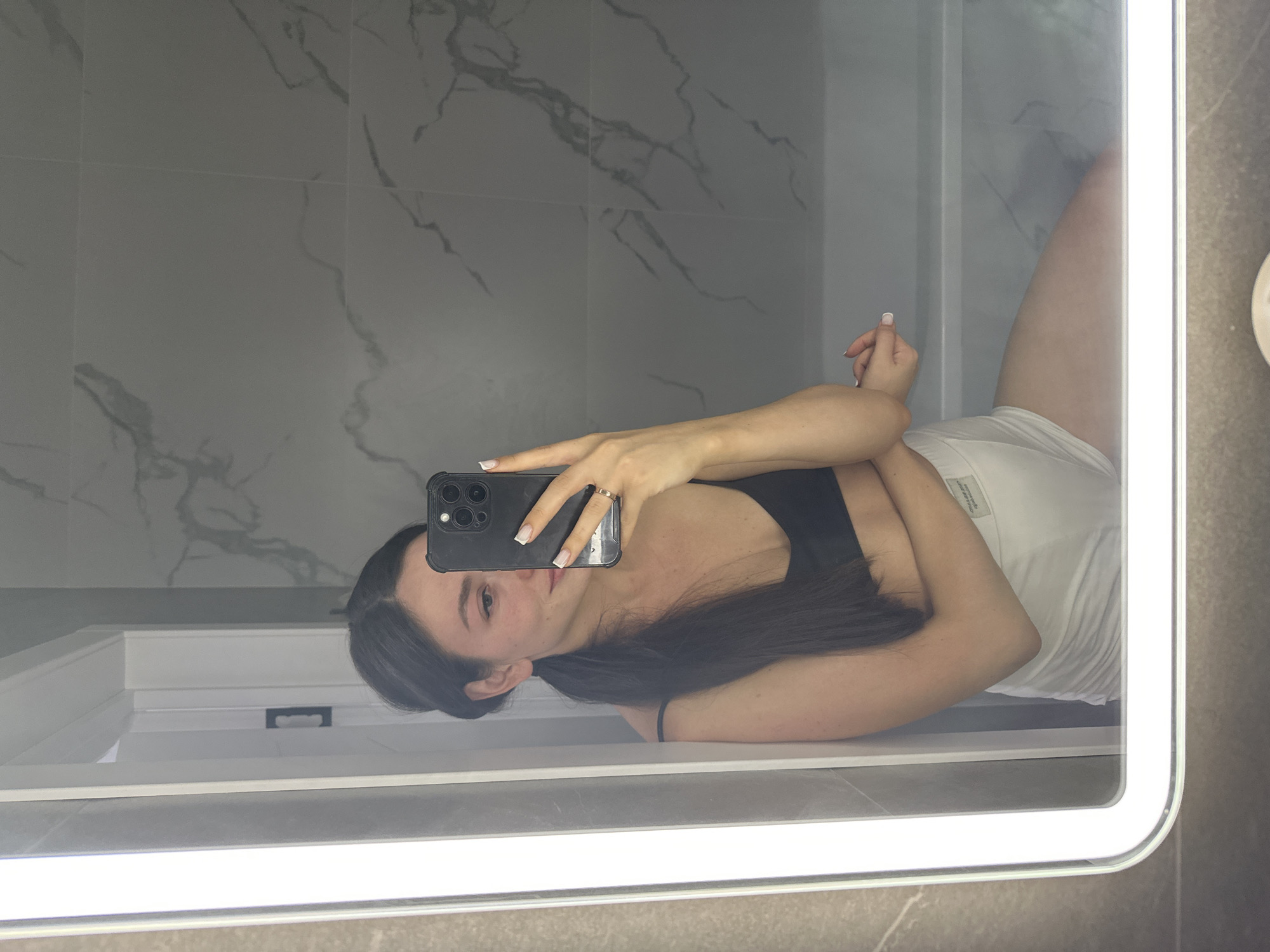 A woman is taking a mirror selfie in a bathroom. She has dark hair pulled back into a ponytail and is wearing a black bra and white shorts. She is holding a black smartphone in her right hand, angled to capture her reflection. The bathroom features marble tile walls and a bright, illuminated mirror. The woman appears to be leaning against a doorframe, with her left arm crossed over her chest and her right arm extended to hold the phone. The reflection shows her face, upper body, and legs. There is no visible indication of the city of Estachi, Russia.