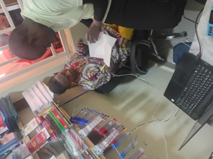 This image depicts an indoor commercial or office setting in Kwazailewa, Nigeria, characterized by stationery supplies. Two adult males are engaged in a transaction or consultation, while a third individual is partially visible in the background.
The male on the left is seated in a black office chair, wearing a multicolored patterned long-sleeved shirt. He holds a yellow pen or marker in his right hand, looking intently at a stack of white papers held by the second male, with a slight smile. The male on the right is standing or leaning, dressed in a light green short-sleeved top worn over a dark long-sleeved undershirt. He holds a stack of white papers, appearing to present or review them with the seated individual, his right hand positioned over the documents.
In the foreground, a black laptop is open on a reflective surface, with a red USB drive or connector inserted into its right side. A light blue keypad mobile phone is positioned to the right of the laptop, connected by a white cable.
To the left, a multi-tiered wooden shelving unit displays various stationery items. Visible products include packages of SanDisk memory cards, stacks of notebooks, and numerous pens of assorted colors (blue, red, black) hanging from hooks. The walls are light-colored. Several white electrical cables are visible, traversing the wall and floor. In the upper right background, a white-barred window or partition reveals the upper portion of a third individual, partially obscured, wearing what appears to be a light pink head covering.