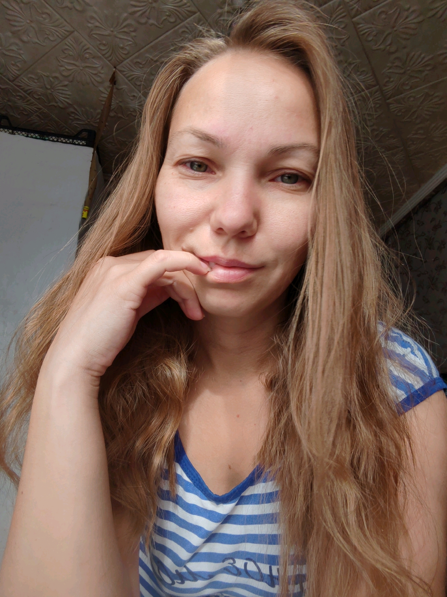 A woman with long, blonde hair and green eyes is looking at the camera. She is wearing a blue and white striped shirt. Her right hand is touching her chin. The background shows a textured ceiling and a wall with a floral pattern.